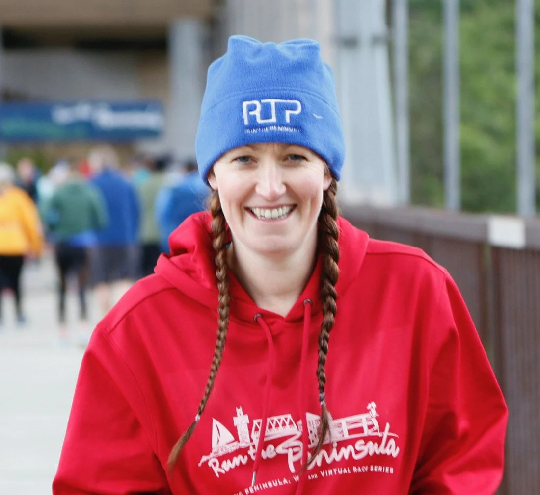 A smiling woman with braided hair wearing a blue beanie and red hoodie at an outdoor event with blurred people in the background.