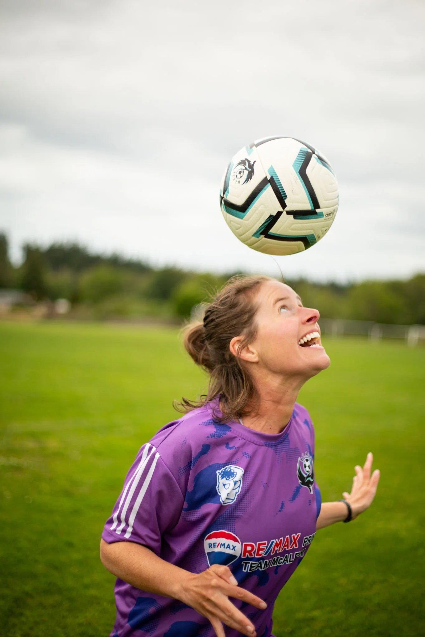 A woman with brown hair in a ponytail, smiling and looking up, wearing a purple soccer jersey, on a grassy field with cloudy sky, heading a soccer ball.