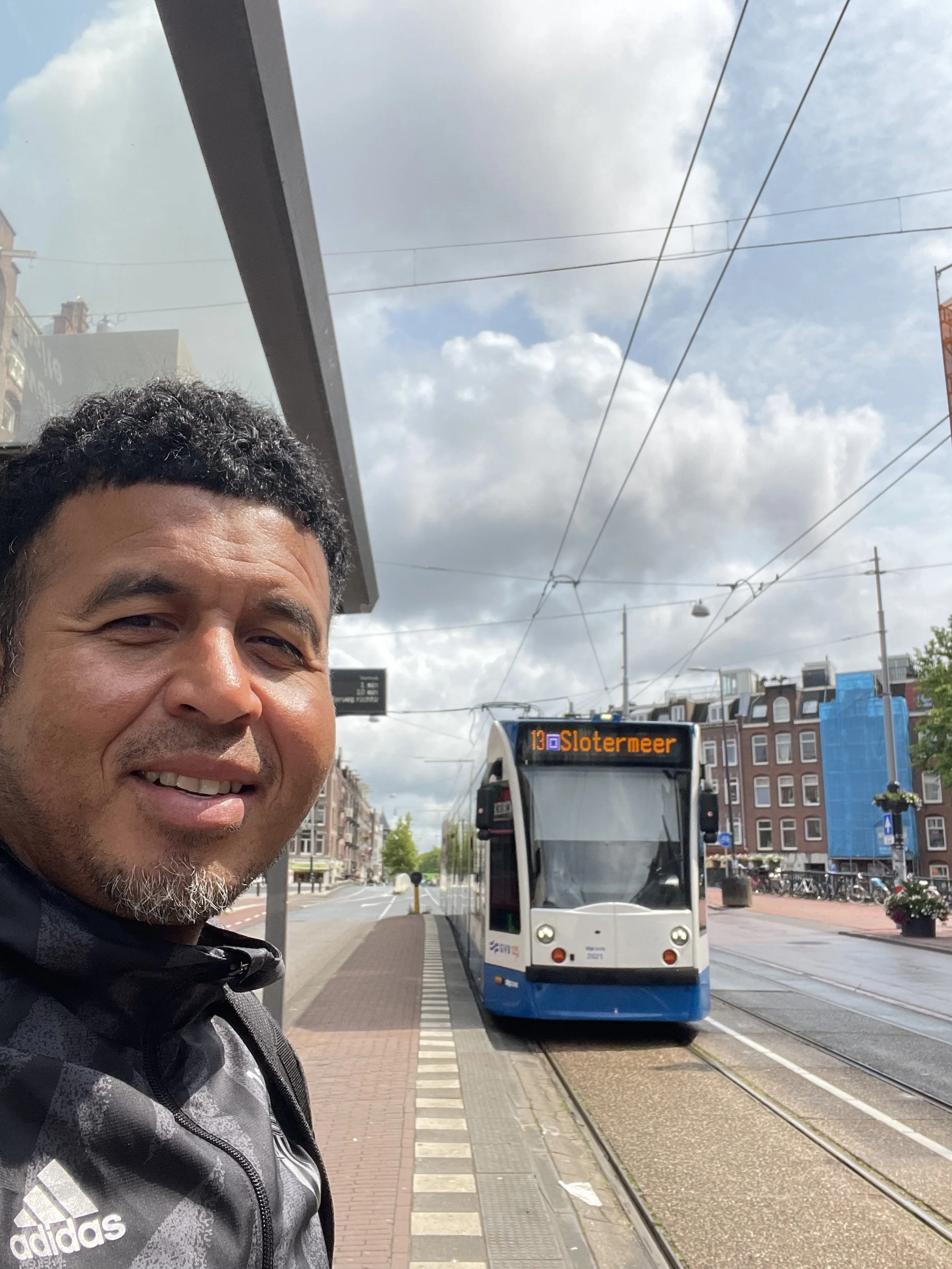 A man smiling at a tram stop with a tram labeled 'Slottermeer' approaching on streetcar tracks. The man is wearing a gray Adidas jacket, and the sky is partly cloudy with buildings and power lines in the background.