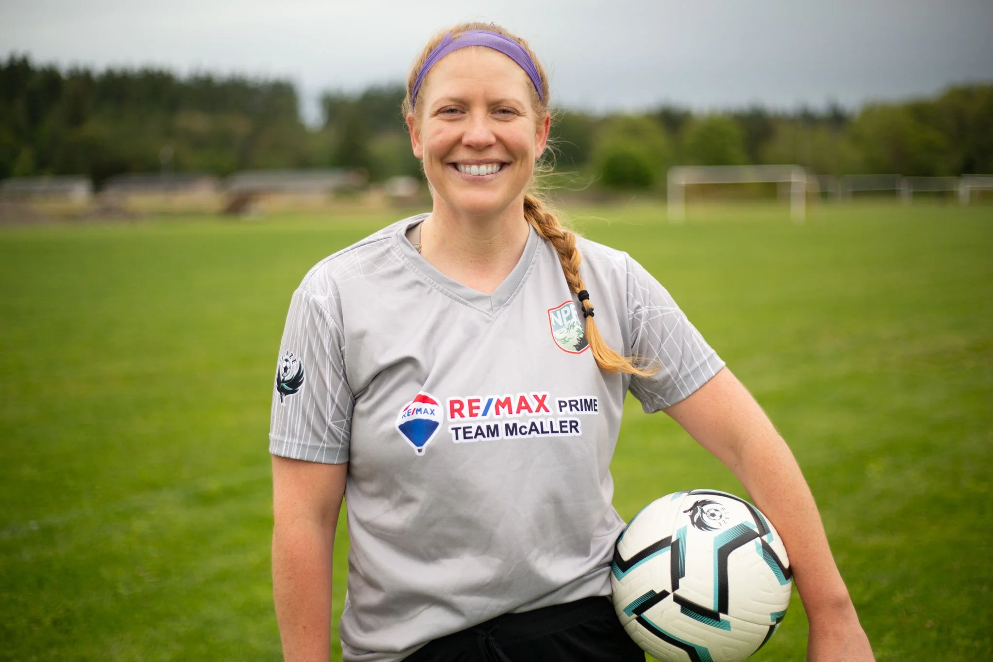 A smiling woman in a gray sports jersey holding a soccer ball outdoors on a grassy field.