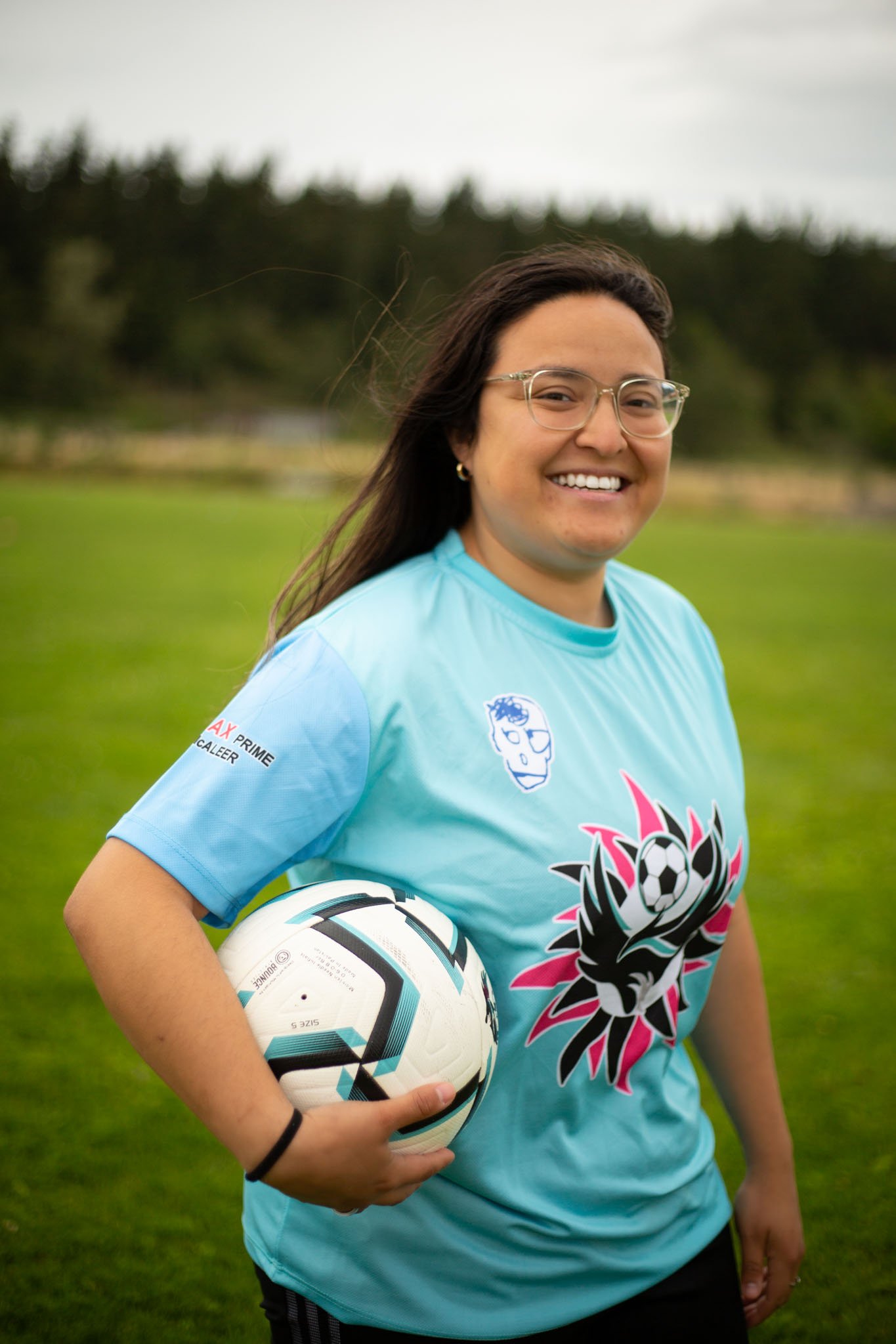 A young woman smiling while holding a soccer ball on a grassy field, wearing glasses and a bright blue soccer jersey with colorful graphics and patches.