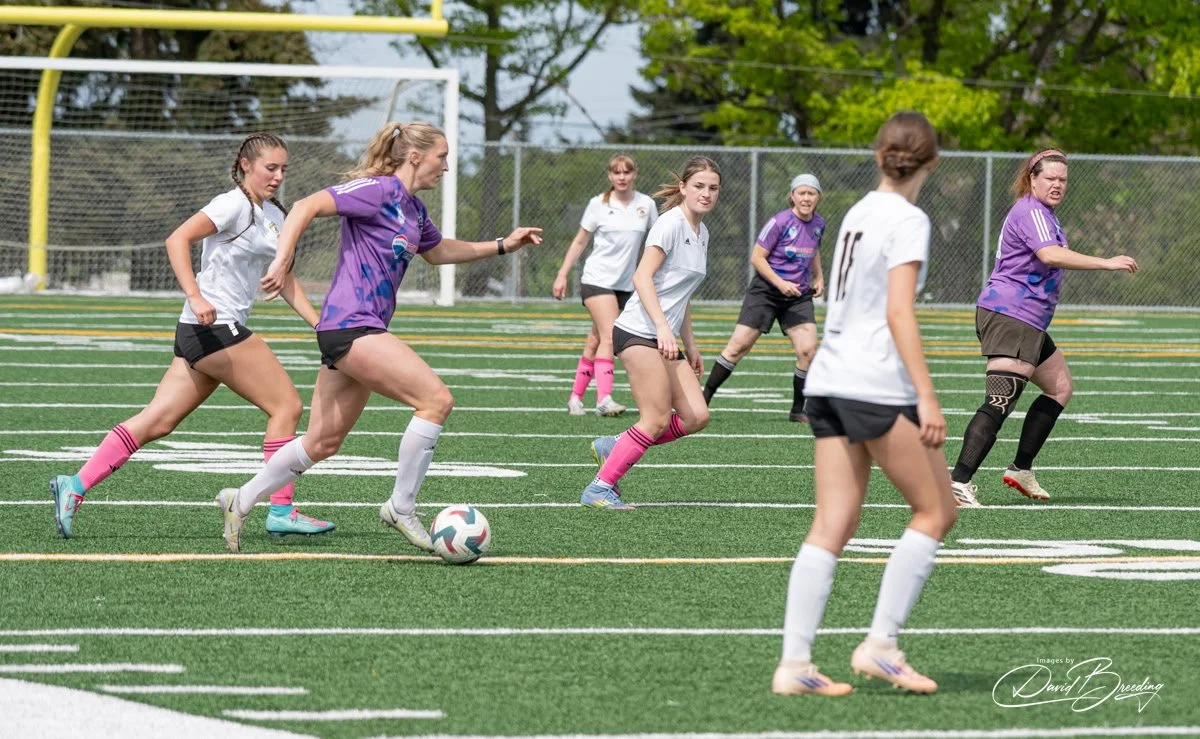 Women's soccer game on a grassy field with players in white and purple jerseys, some wearing pink socks, with a goalpost and chain-link fence in the background.