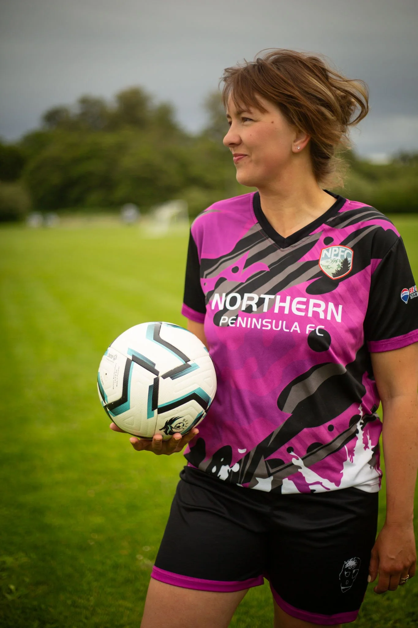 A woman in a pink and black athletic jersey holding a soccer ball on a grassy field, with trees and a cloudy sky in the background.