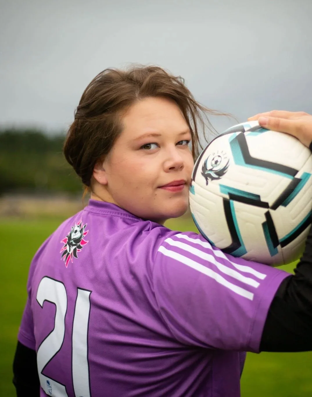 A woman in a purple soccer jersey holding a soccer ball on a grassy field.