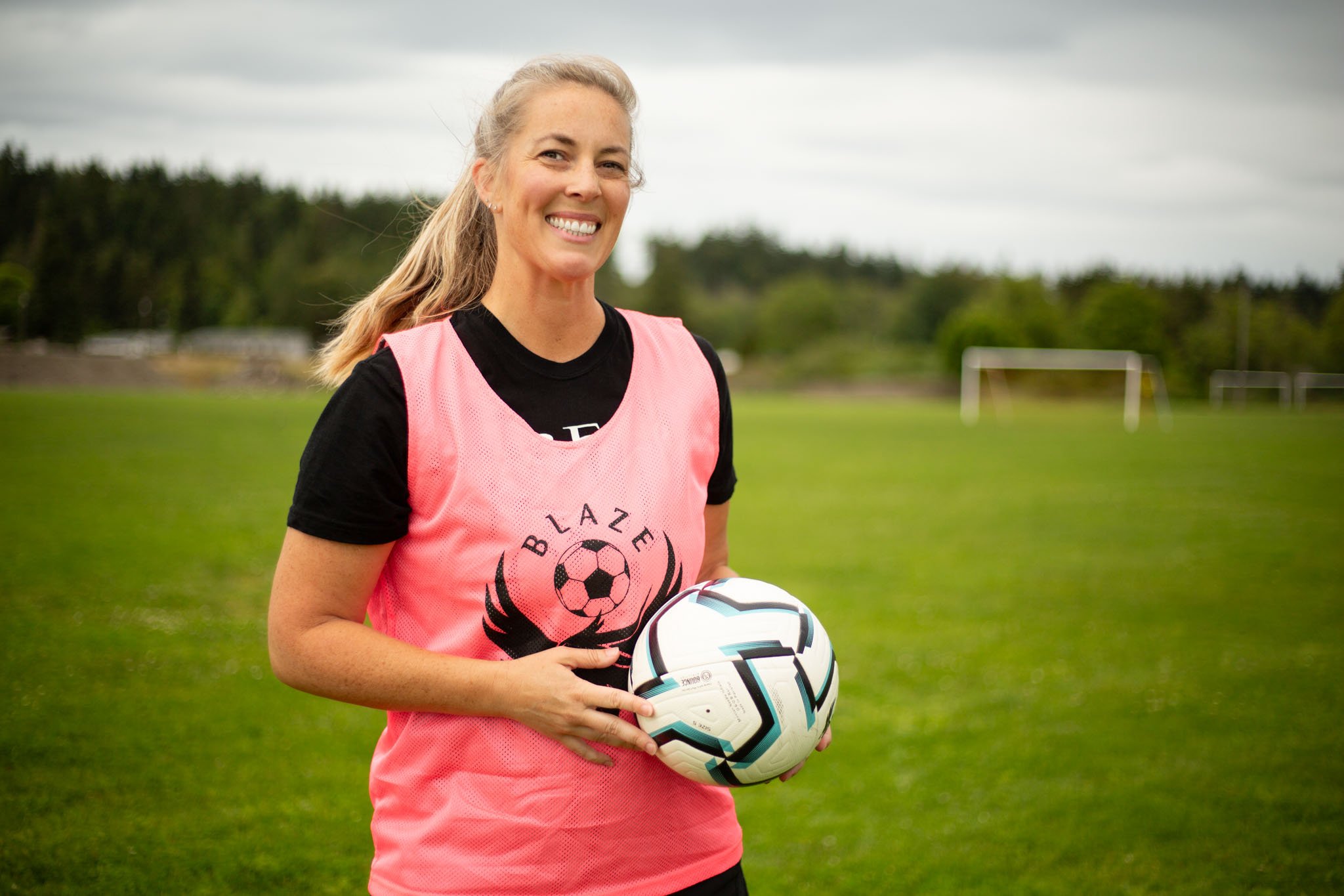 Smiling blonde woman in pink soccer jersey holding a soccer ball on a grassy field with goal posts in the background.