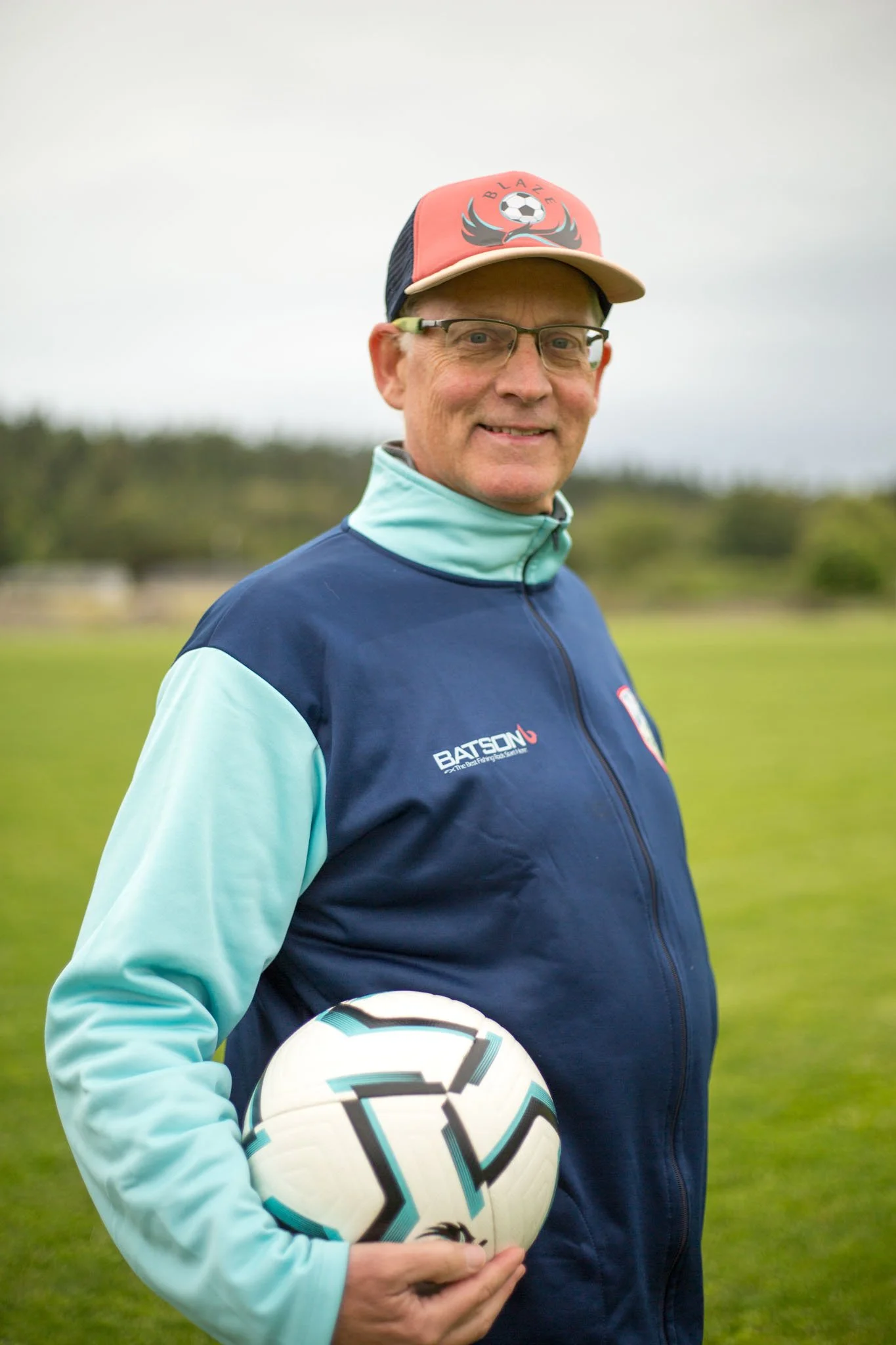 A man wearing glasses, a baseball cap with a soccer ball and flaming wings logo, a light blue jacket, holding a soccer ball, standing outdoors on a grassy field, with a blurred background of trees and overcast sky.