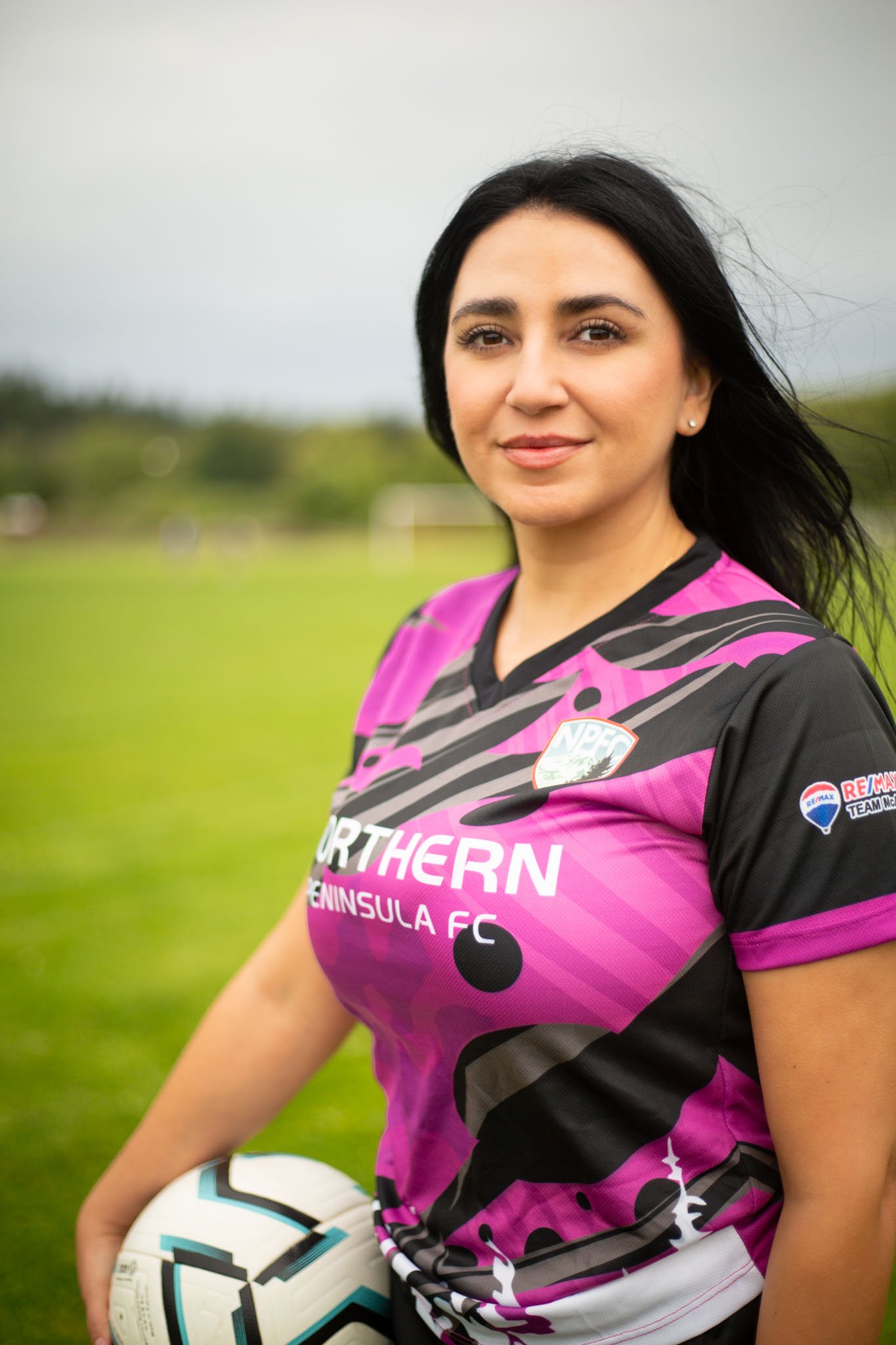 A woman with dark hair and fair skin wearing a pink and black sports jersey with the words 'Northern Peninsula FC' on it, holding a white soccer ball with black and teal accents, standing on a grassy field during daytime.