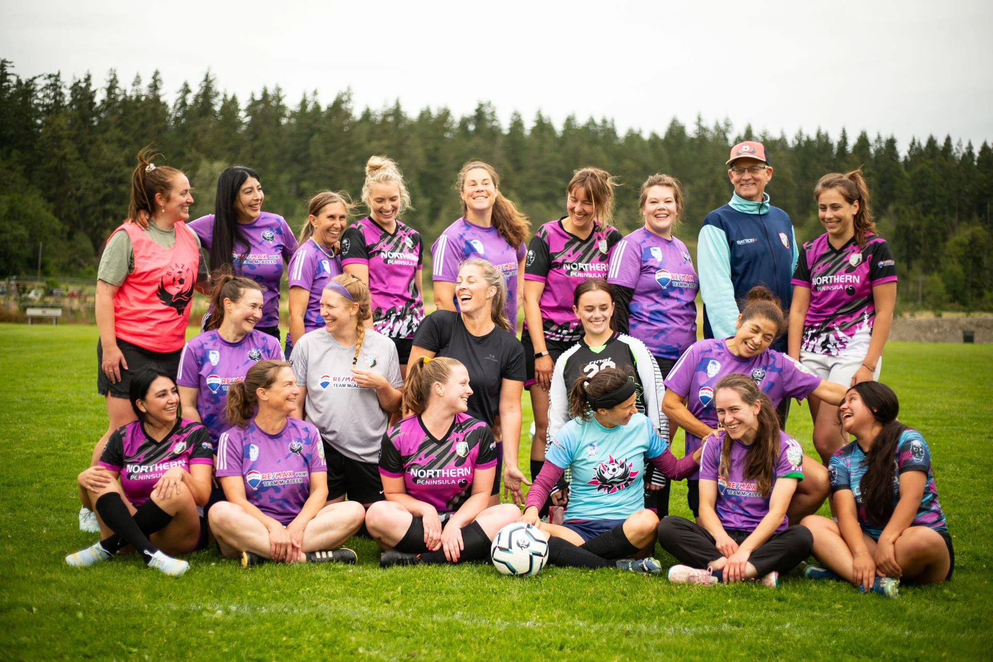 Group of female soccer players and coach posing on a grassy field during daytime, with trees in the background.