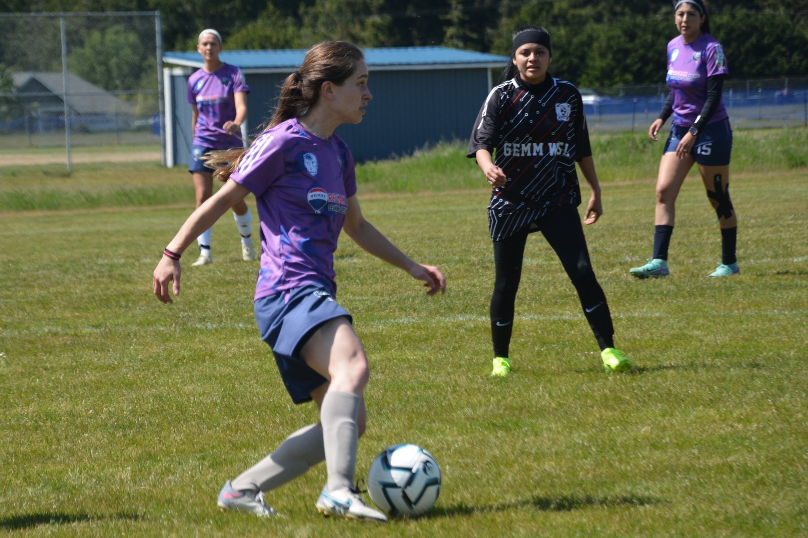 Four women playing soccer on a grassy field during daytime, with two in purple jerseys and two in black jerseys, with trees and a fence in the background.
