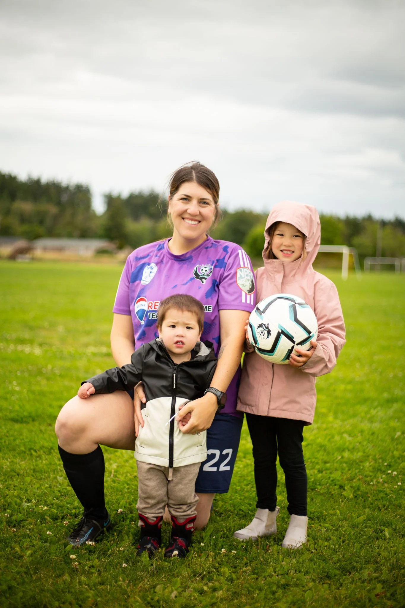 A woman in a purple sports jersey and two children standing on a grassy field. The woman is smiling at the camera, and one child in a pink hooded jacket is holding a soccer ball. The other child, in a black jacket, is standing close to the woman. The background shows trees and a cloudy sky.