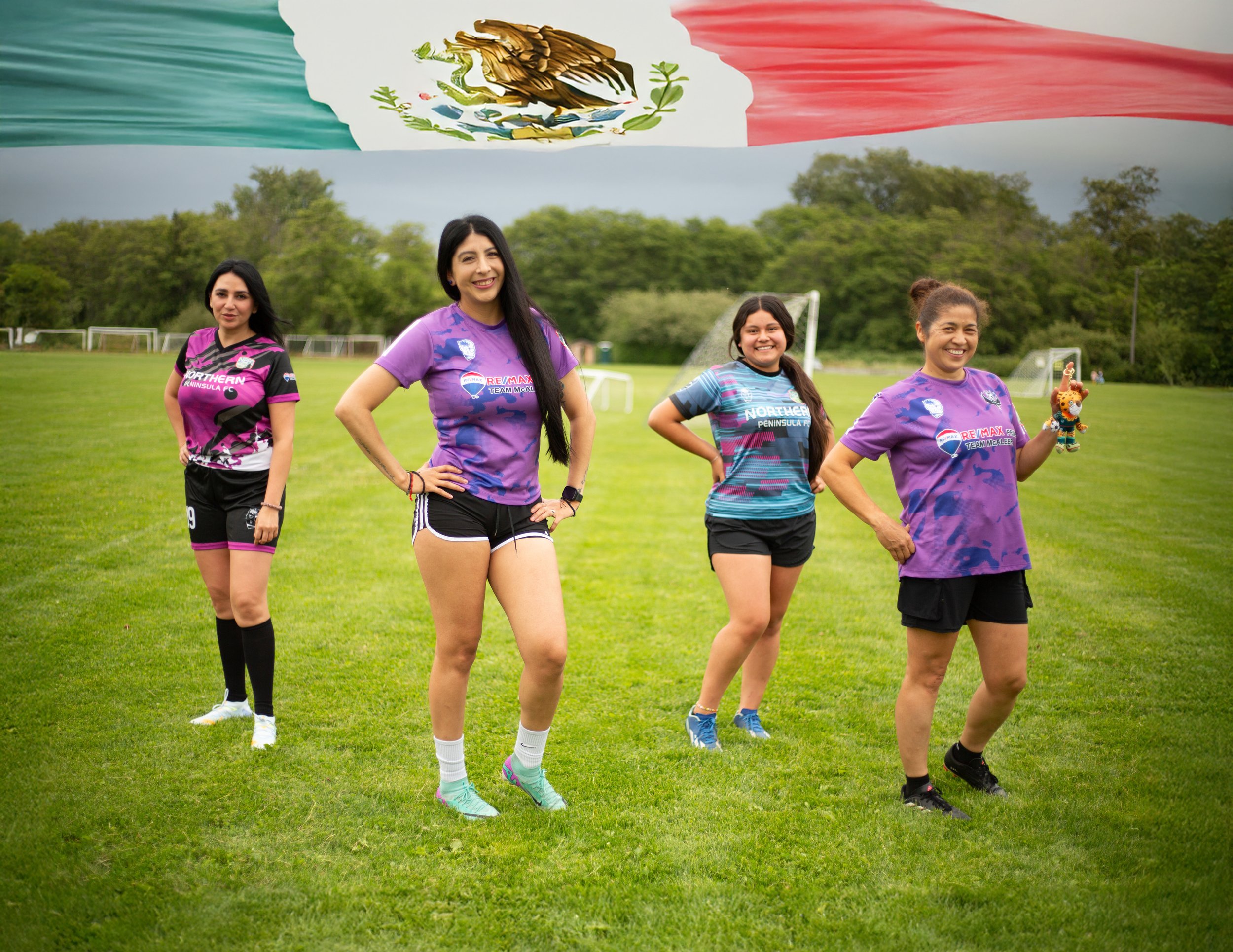 Group of women standing outdoors on a grassy field, smiling and wearing sports jerseys, with a large flag featuring the Mexican coat of arms and green, white, and red stripes in the background.