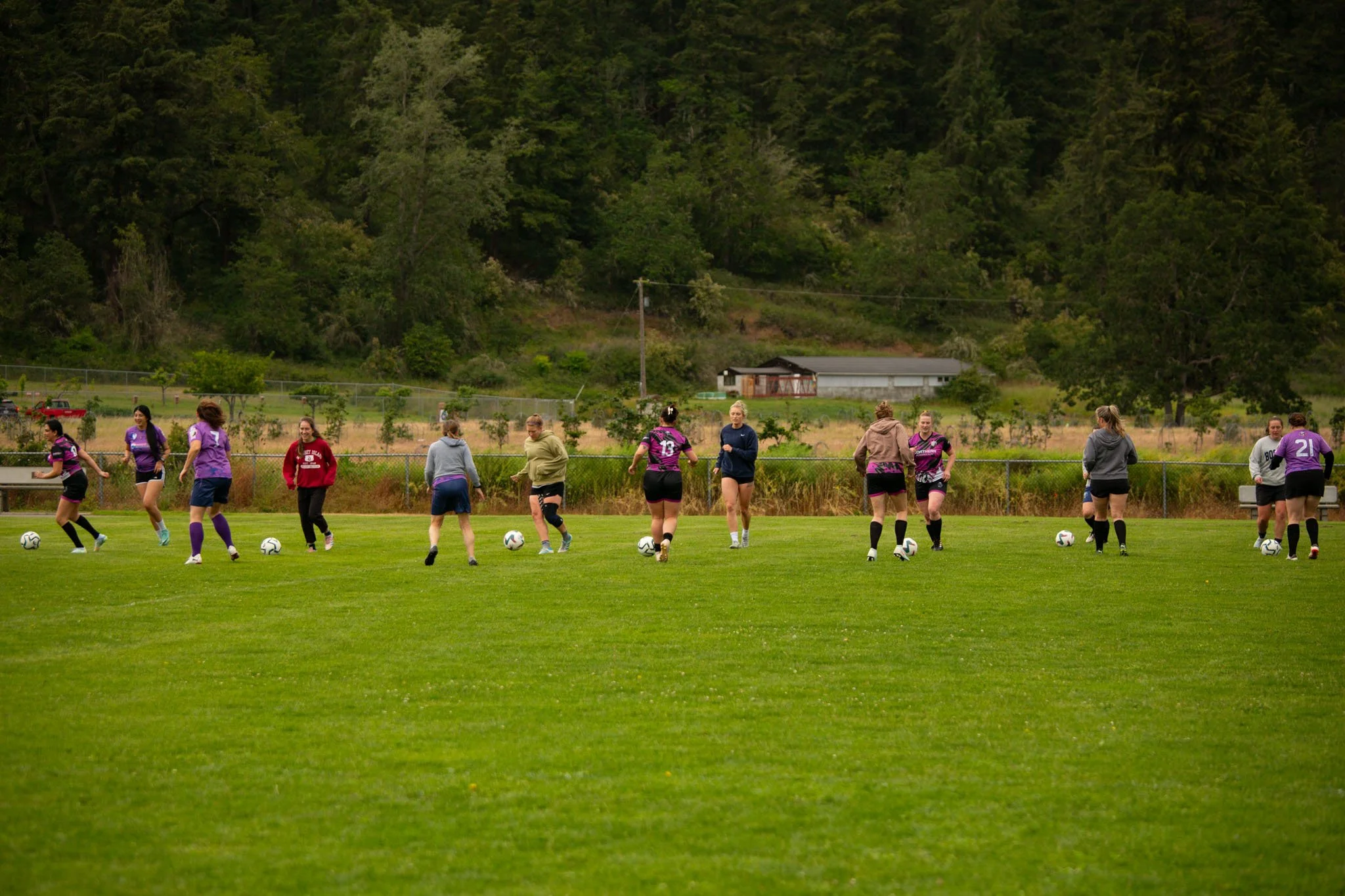 A group of women playing soccer on a grassy field, with a backdrop of trees and a small building.