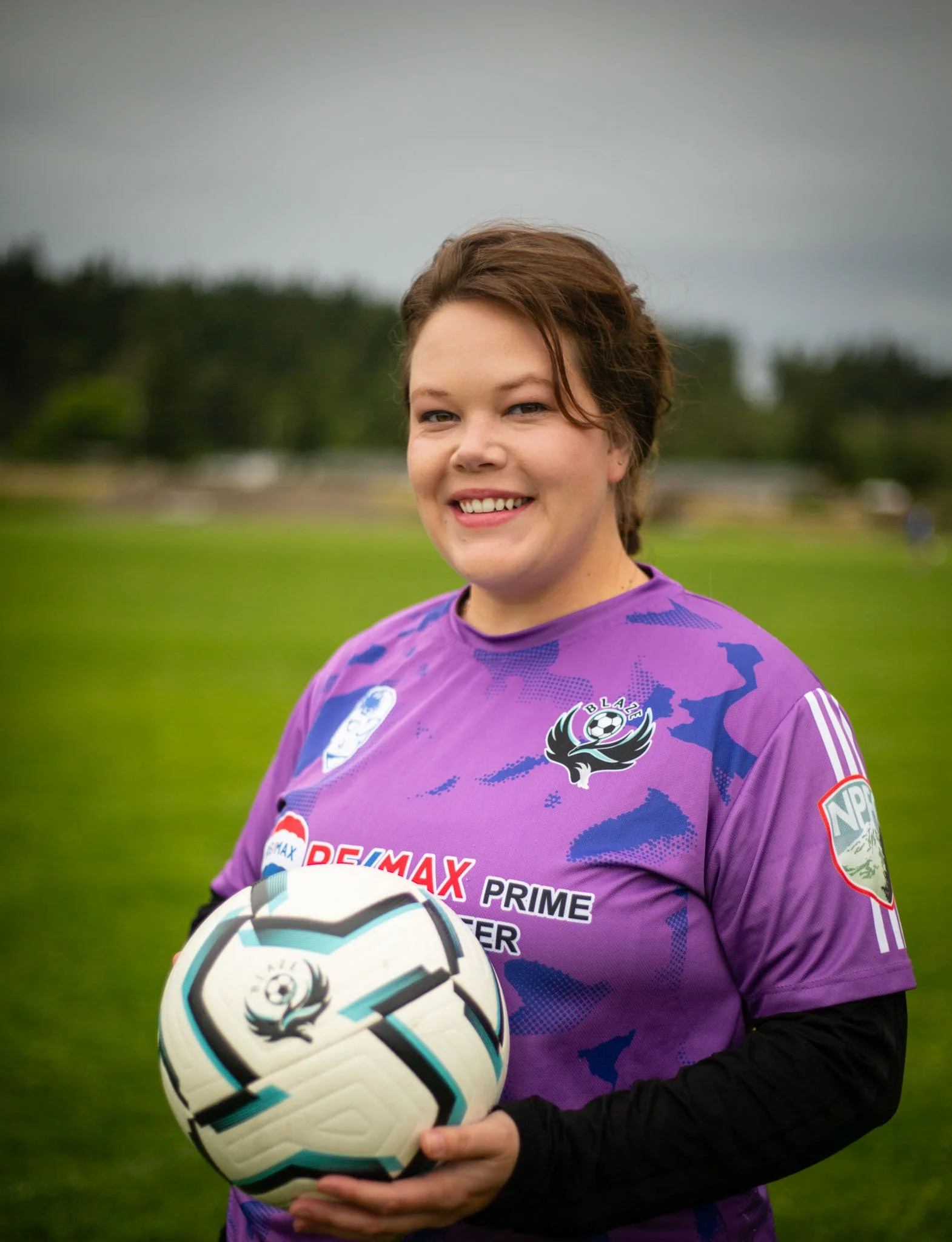A woman with short brown hair smiling while holding a soccer ball outdoors on a grassy field, wearing a purple sports jersey with various sponsor logos.