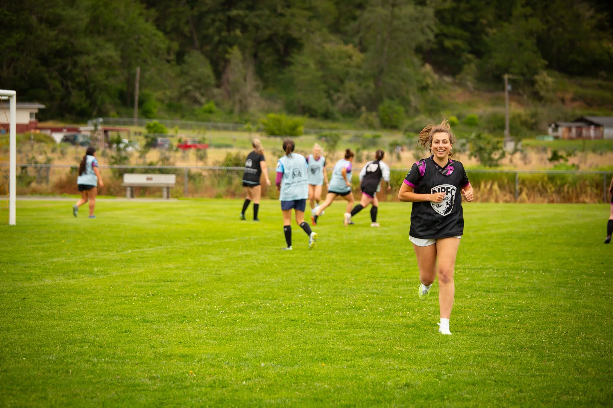 A group of young women playing soccer on a grassy field, with one smiling woman running toward the camera, and others in the background near a goalpost.