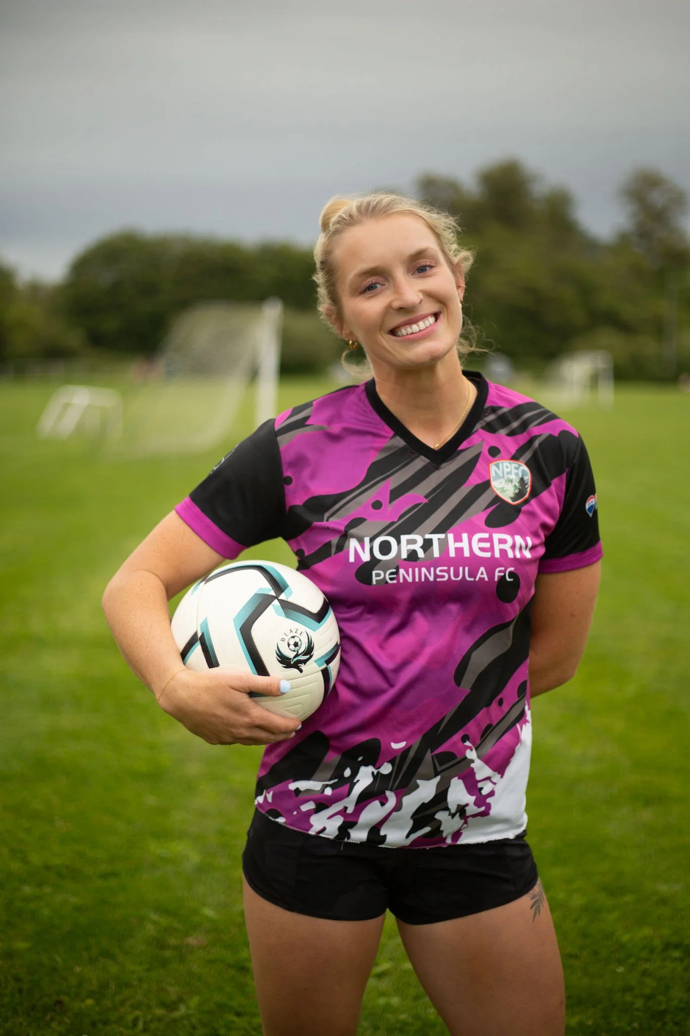 A smiling young woman in a pink and black sports jersey holding a soccer ball on a grassy field with soccer goals in the background.