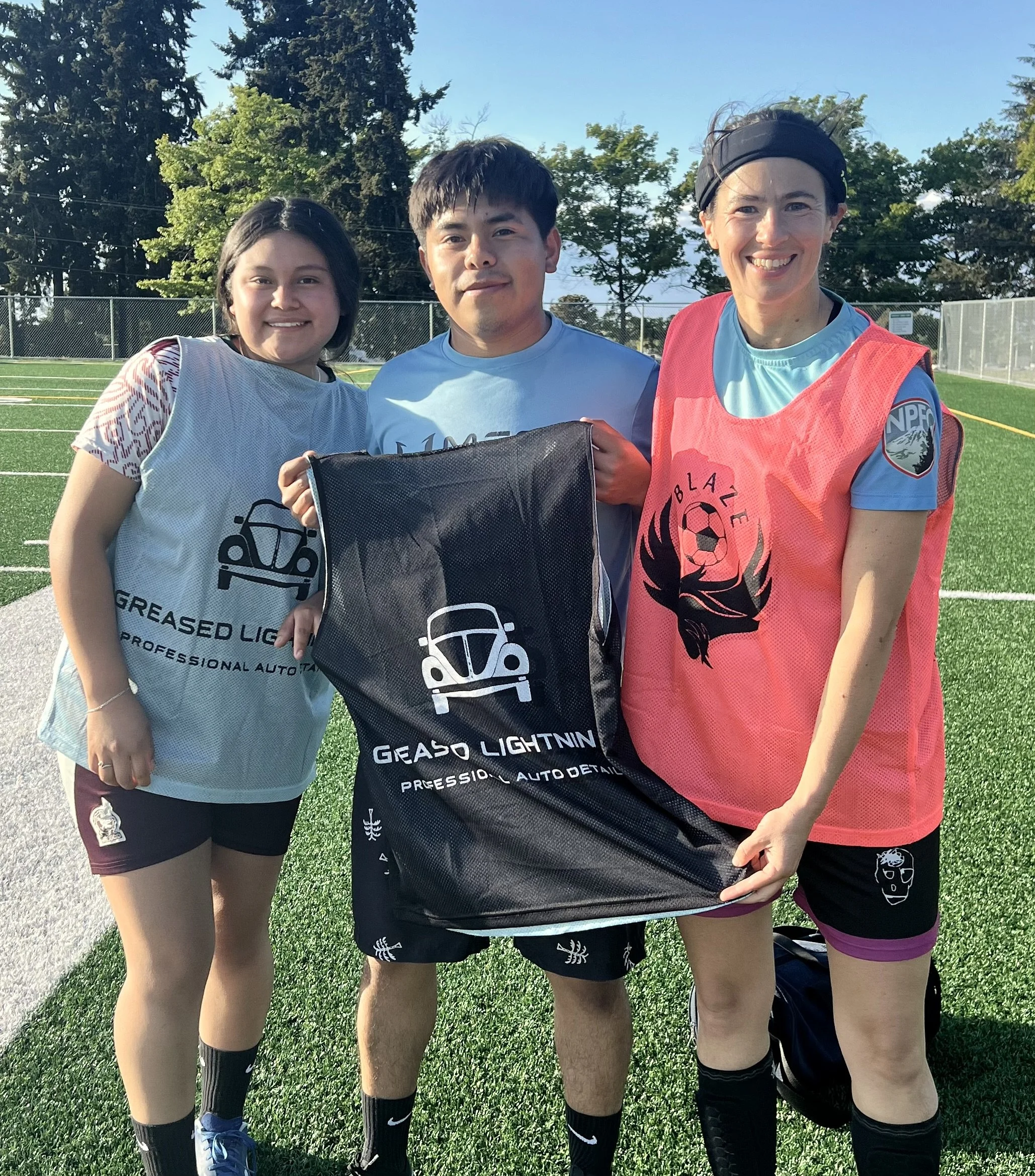 Three young women and one young man standing on a grassy field, posing for a photo. They are dressed in athletic wear, some with sports vests and others with shorts and t-shirts. They are smiling and standing close together, with trees and a chain-link fence in the background under a clear blue sky.