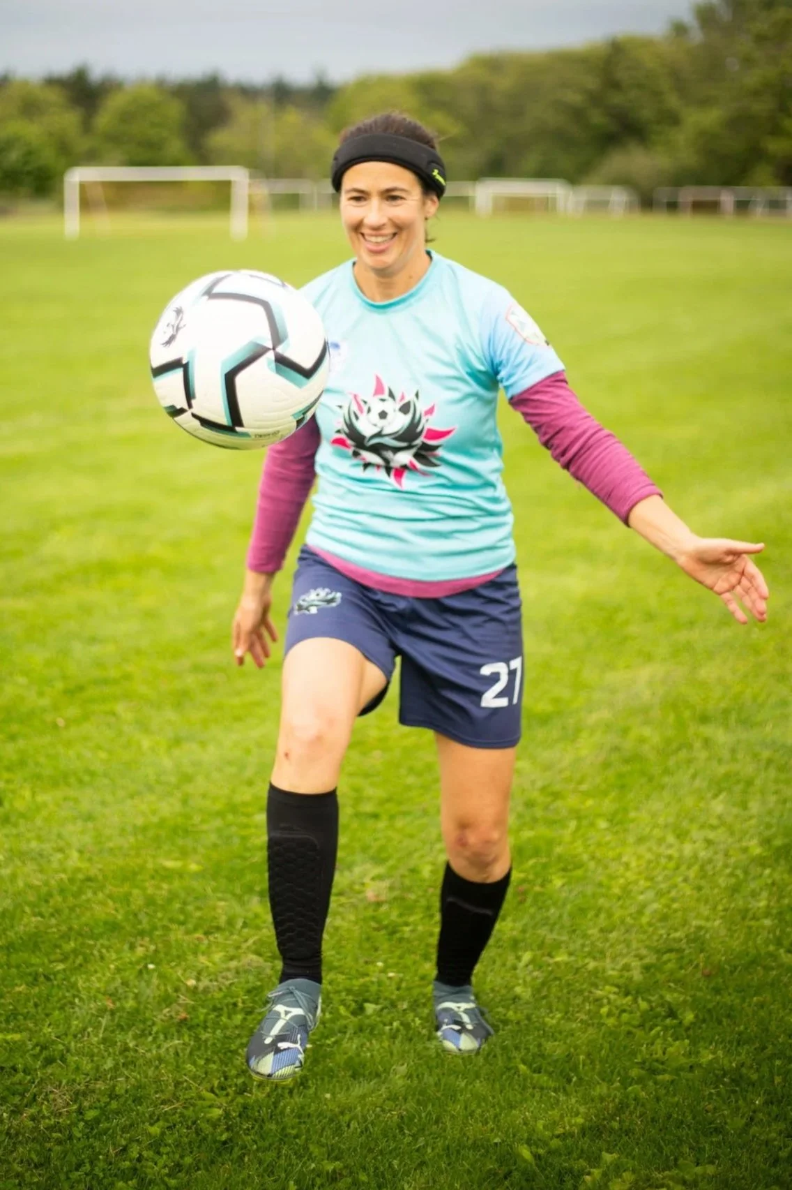 A woman on a grassy field. She's playing soccer, wearing a black headband, a light blue jersey with a panda graphic, navy shorts with the number 27, long black socks, and athletic shoes. She is smiling and appears to be about to kick or control a soccer ball.
