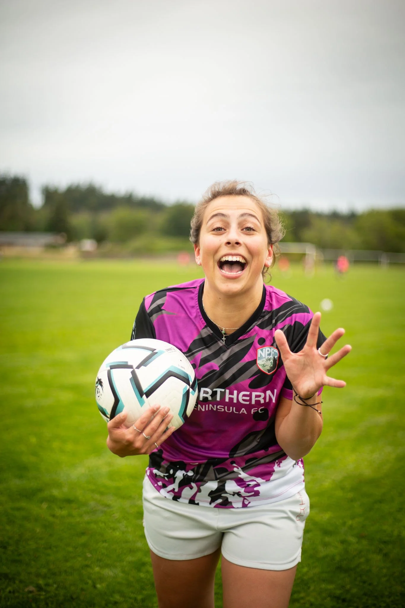 A woman wearing a black and purple sports jersey with the text 'Northern Peninsula FC,' standing on a grassy sports field, holding a soccer ball, and smiling with her mouth open. She is making a peace sign with her right hand, and there are players and goalposts visible in the background.