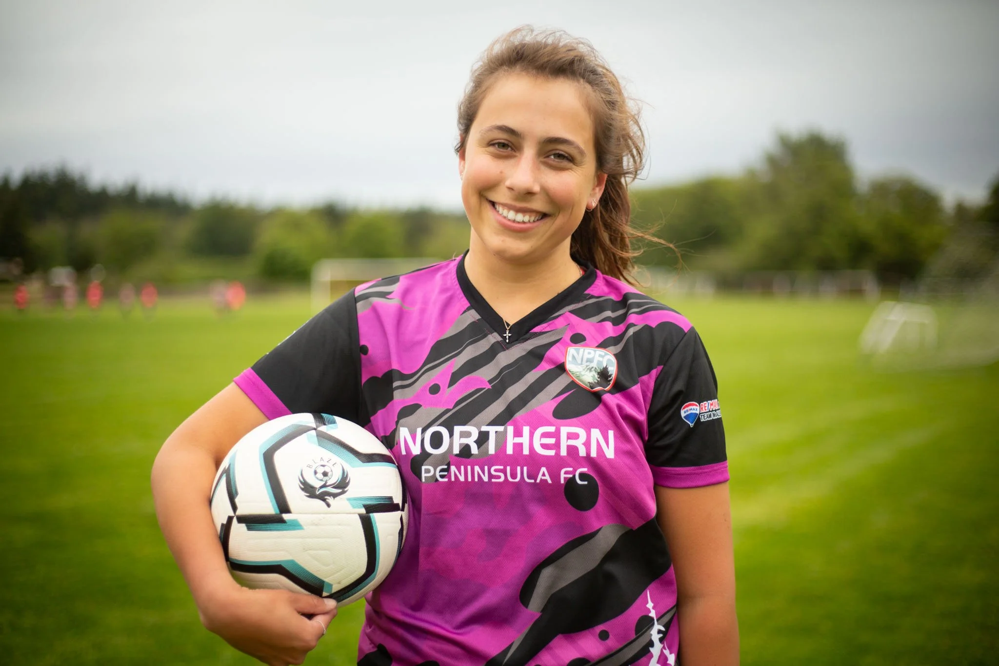 A young female soccer player holding a soccer ball, wearing a black and pink jersey with the logo of Northern Peninsula FC, standing on a grassy field with trees and other players in the background.