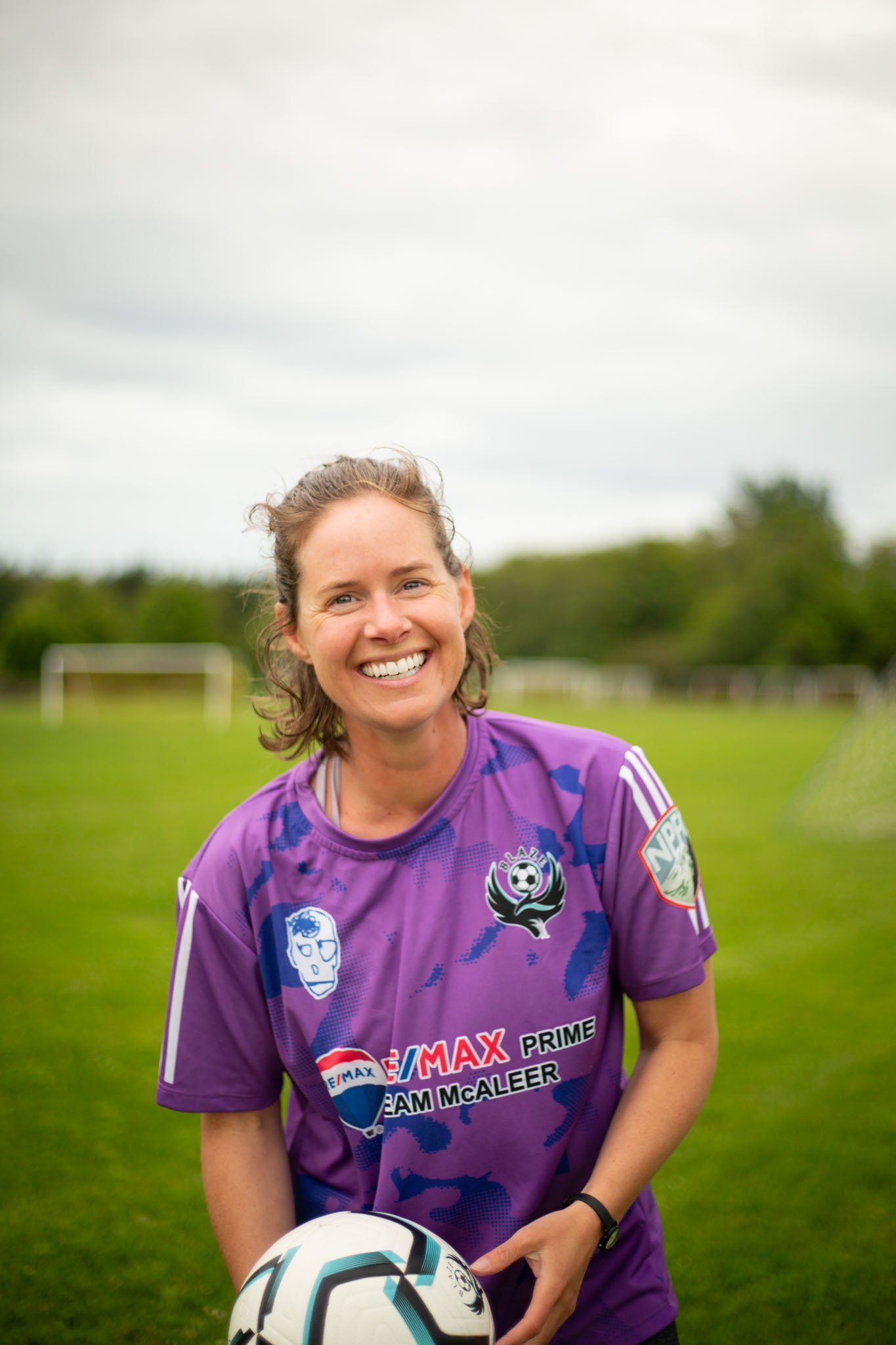 A woman in a purple soccer jersey holding a soccer ball on a field with goal posts in the background.