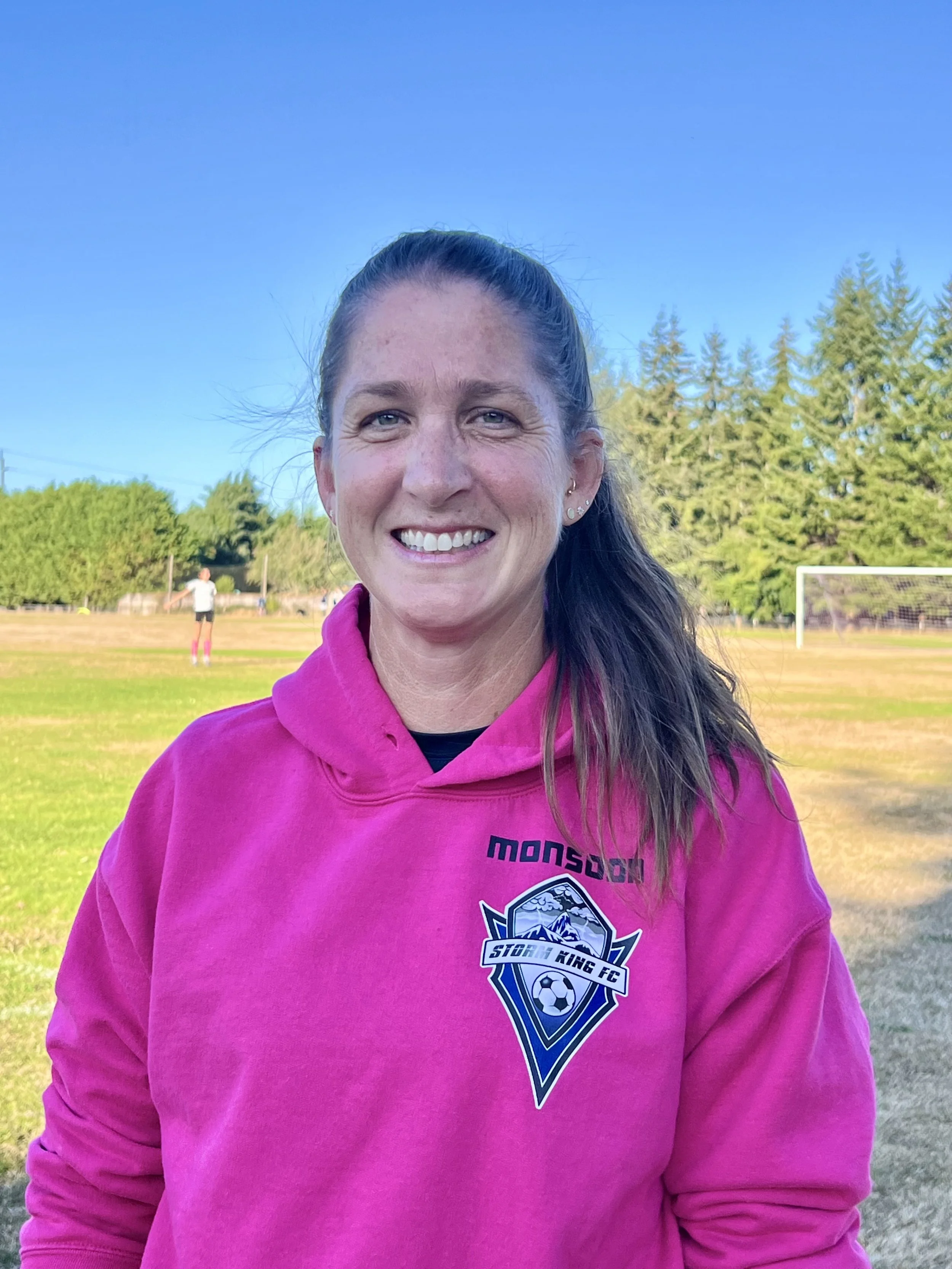 A woman with long dark hair wearing a bright pink hoodie with a sports team logo, standing on a field with trees and a clear blue sky in the background. There is another person playing soccer in the distance.