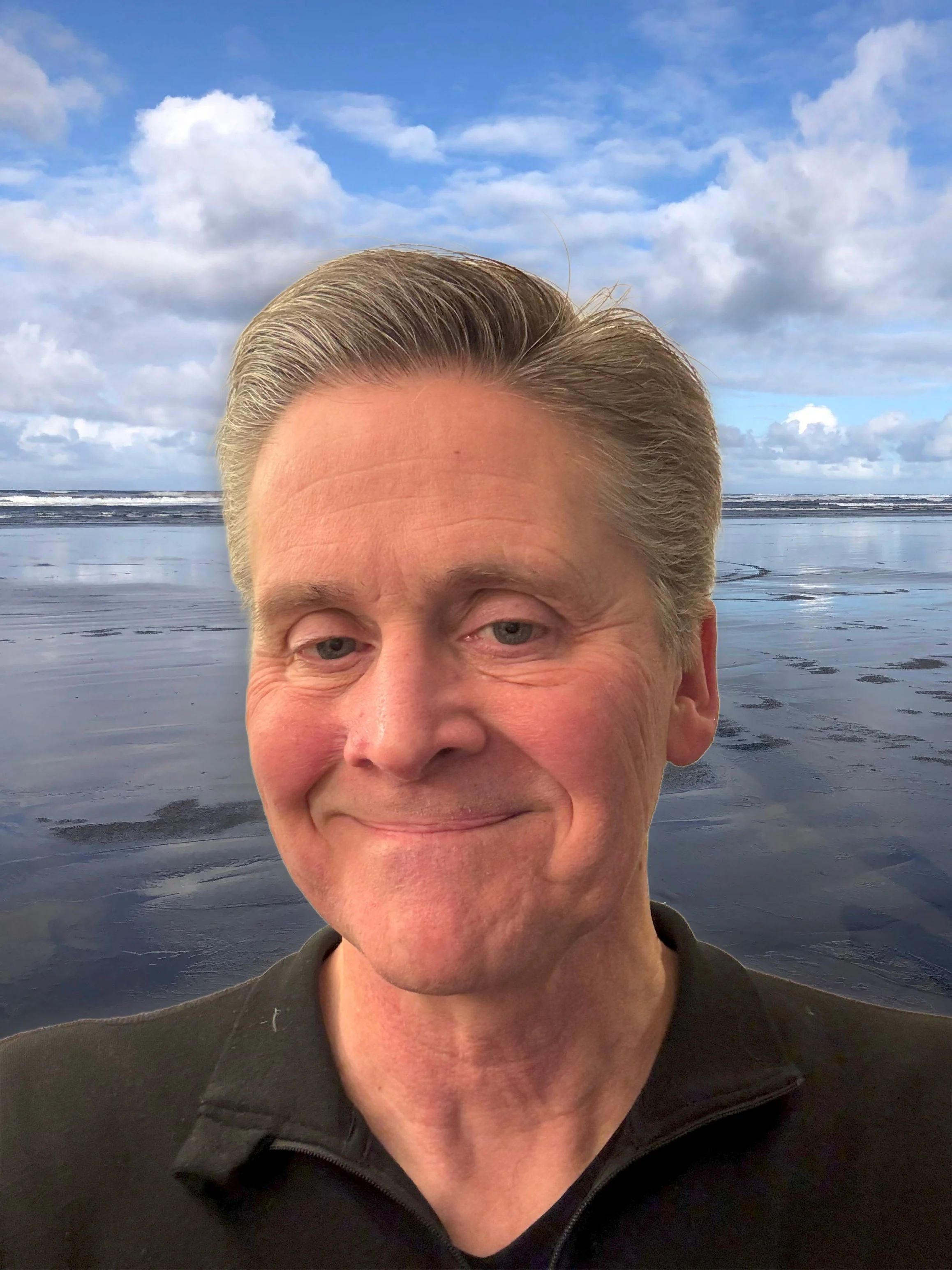 A man with light-colored hair smiling in front of a beach with an overcast sky and ocean waves.