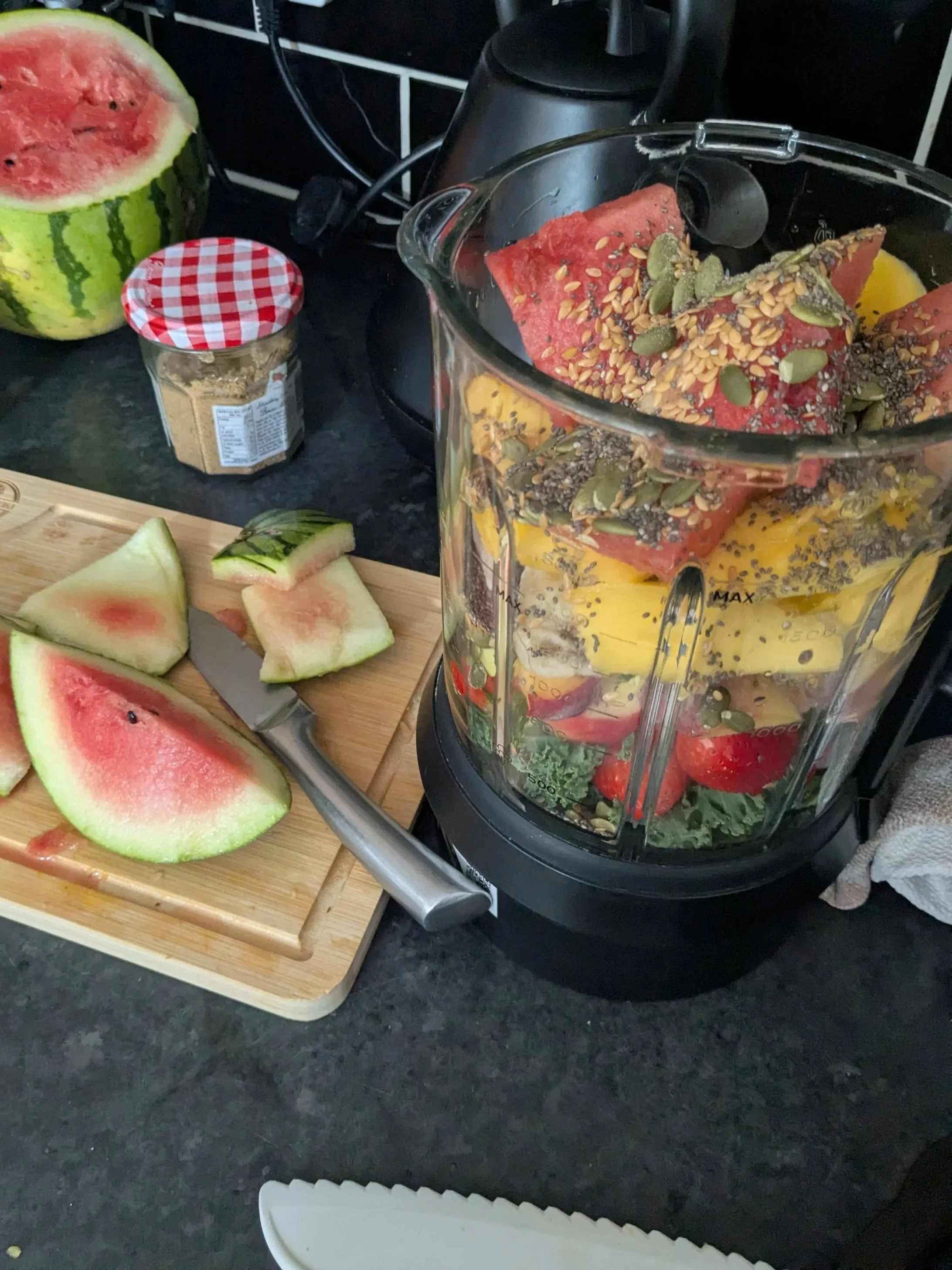 A blender filled with watermelon slices and pumpkin seeds, a wooden cutting board with watermelon slices and a knife, a jar of peanut butter with red and white checkered lid, and a half watermelon on a black countertop.