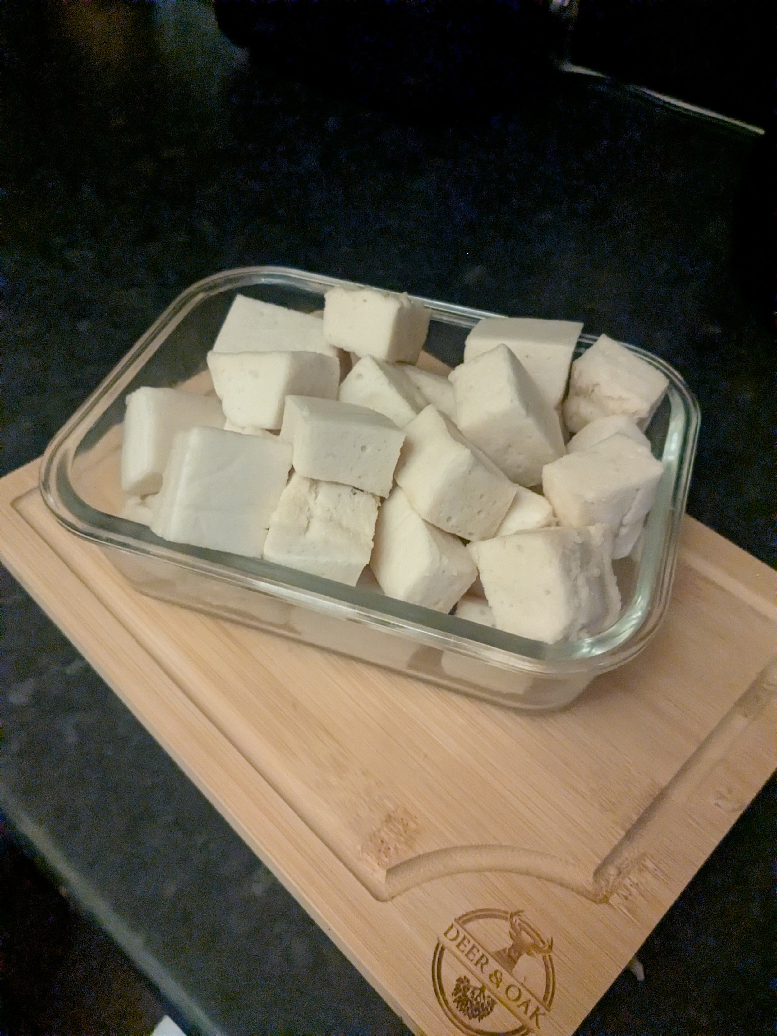 A glass container filled with cubed pieces of tofu placed on a wooden cutting board on a dark surface.