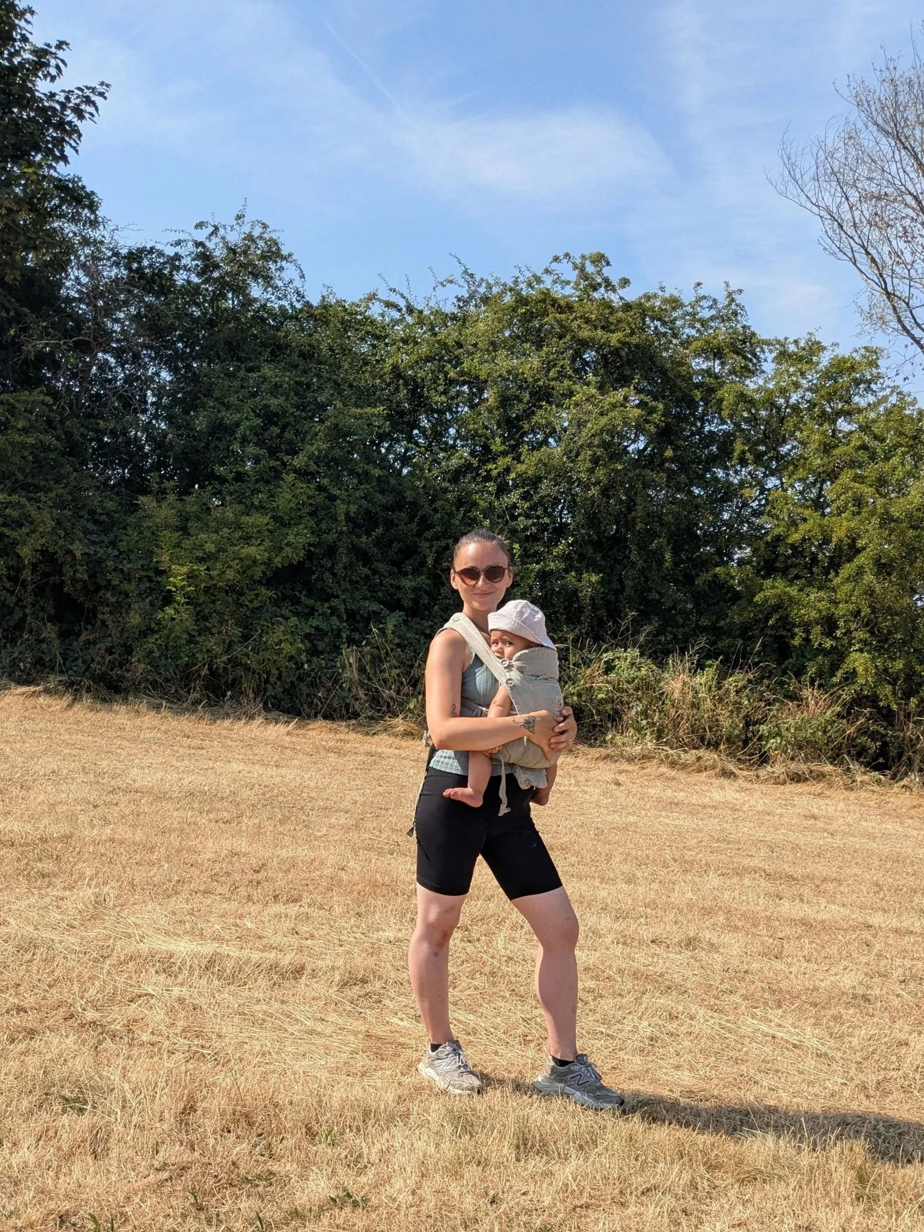 A woman wearing sunglasses and athletic clothing stands outdoors on a clear day, holding a young child in a baby carrier with a wide-brimmed hat. They are on a dry, grassy field with green trees and a blue sky in the background.