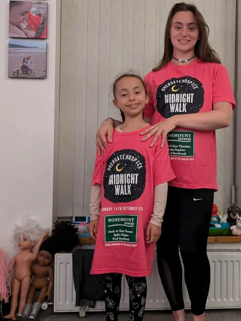 A woman and a young girl are standing indoors, both wearing pink T-shirts with a moon and stars, promoting a midnight walk event. The woman has long brown hair, is smiling, and is standing with her arm around the girl. The girl has dark braided hair 