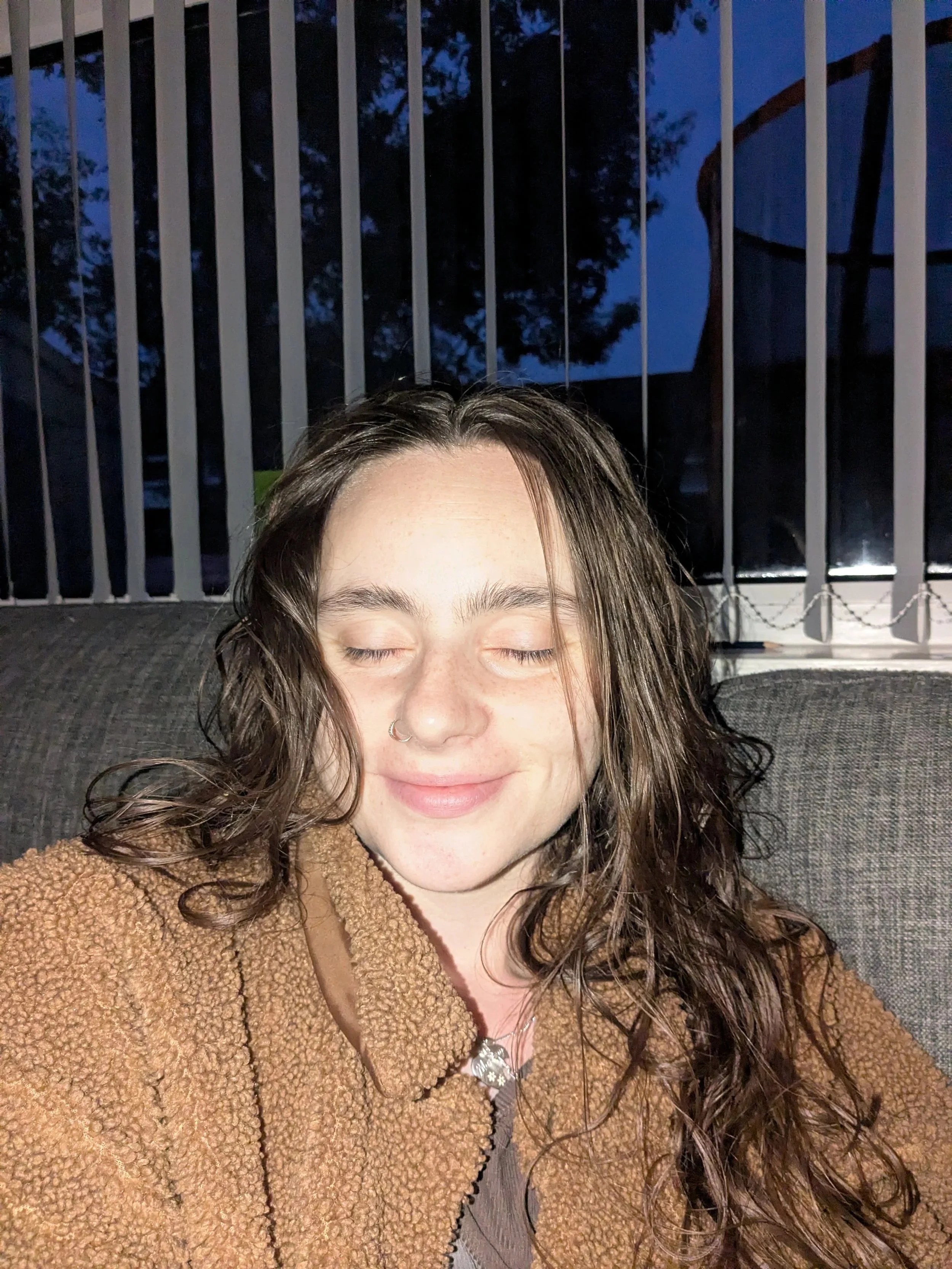 Close-up of a woman with curly brown hair and a nose ring, smiling with eyes closed, wearing a brown fluffy jacket and a necklace with a pendant. The background shows a balcony with a metal fence and trees outside, and it appears to be evening or nig