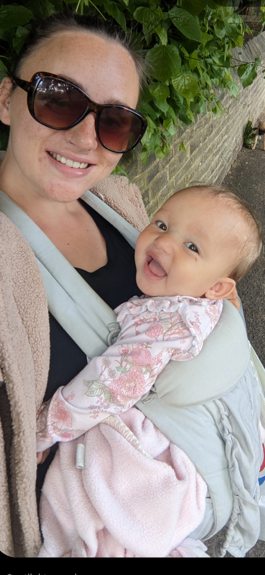 A woman with sunglasses and a nose piercing smiling while holding a laughing baby in a carrier. They are outdoors with green leaves and a brick wall in the background.