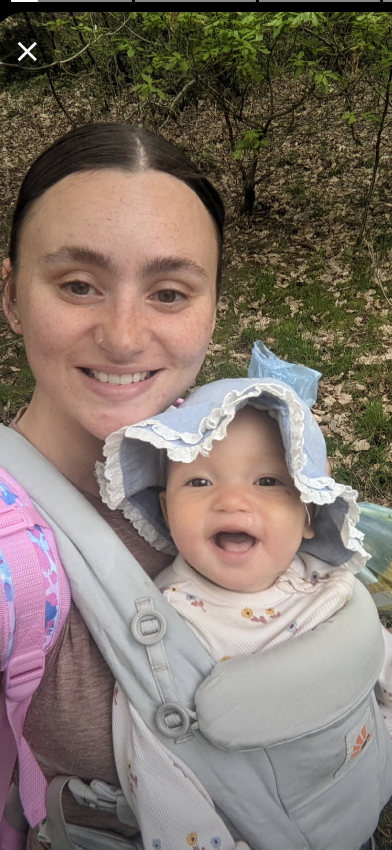 A woman with dark hair and a baby with a sun hat, both smiling, outdoors in a wooded area.