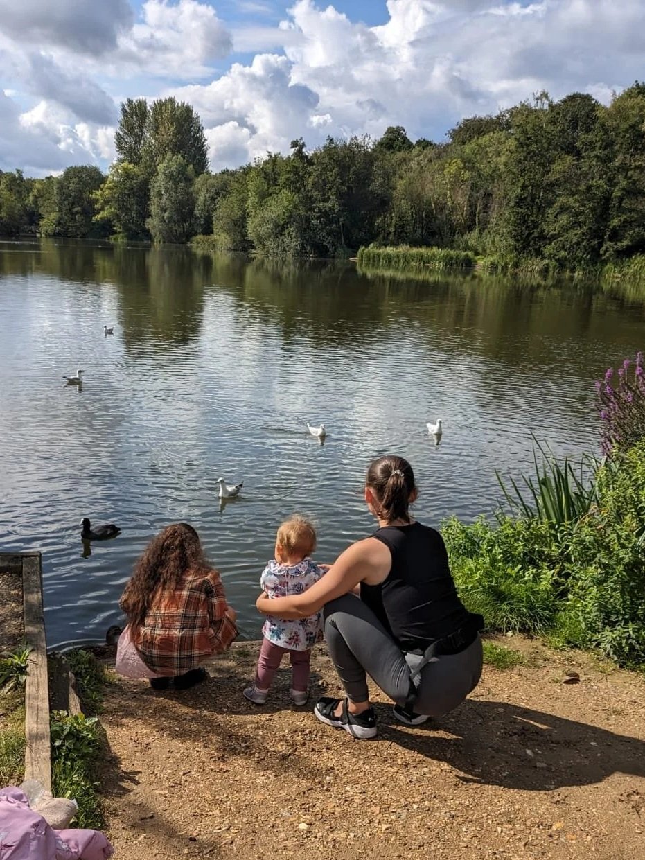 A woman crouches next to a child by a lake, with another child nearby, feeding or observing ducks on the water. They are surrounded by greenery and trees, with a partly cloudy sky overhead.