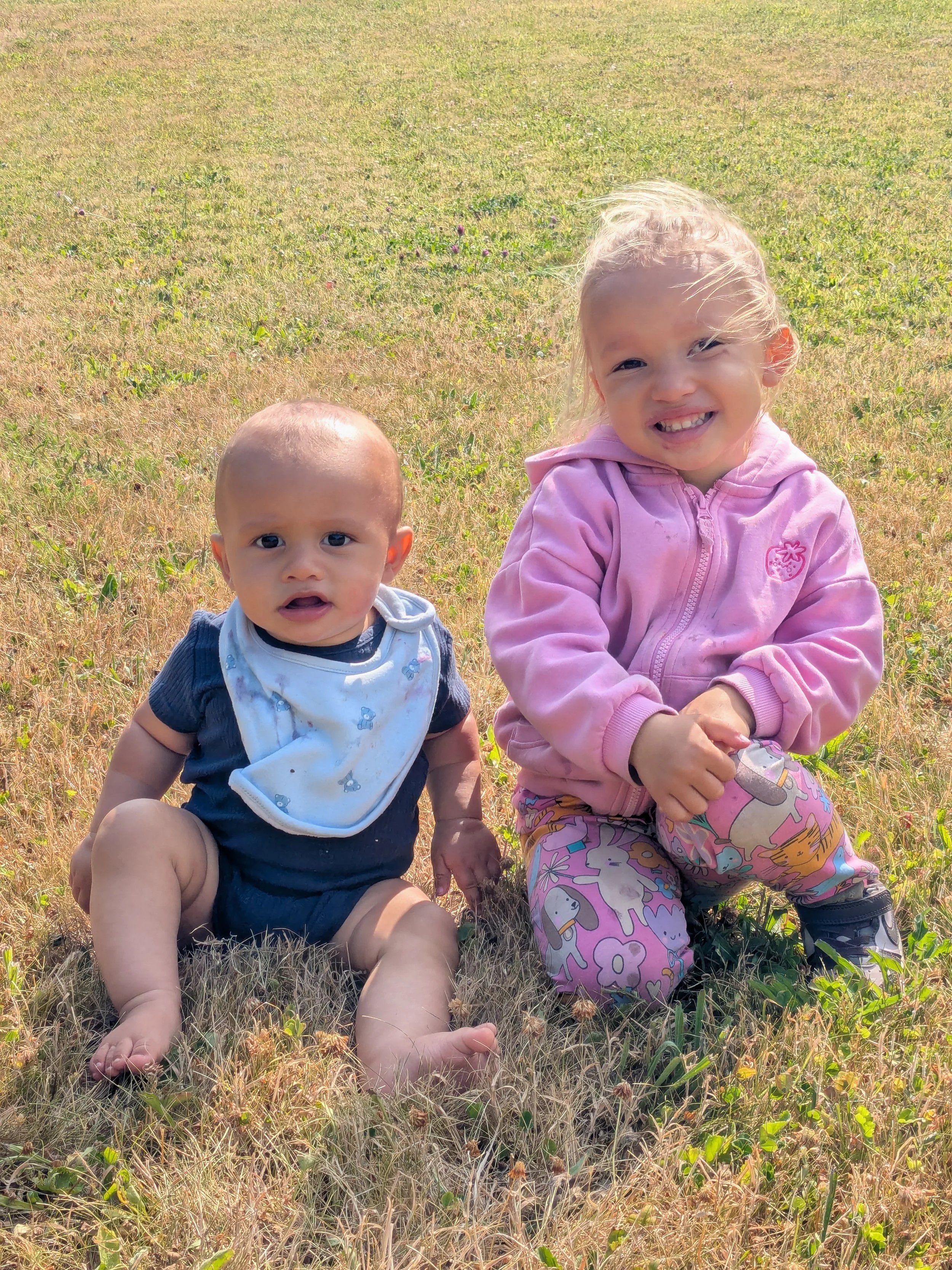 Two young children, a baby boy and a girl, sitting on grass in an open field. The girl is smiling and wearing a pink hoodie, while the baby boy has a neutral expression and is dressed in a dark shirt with a white bib. The background shows a mostly dry grassy field with some small green plants.