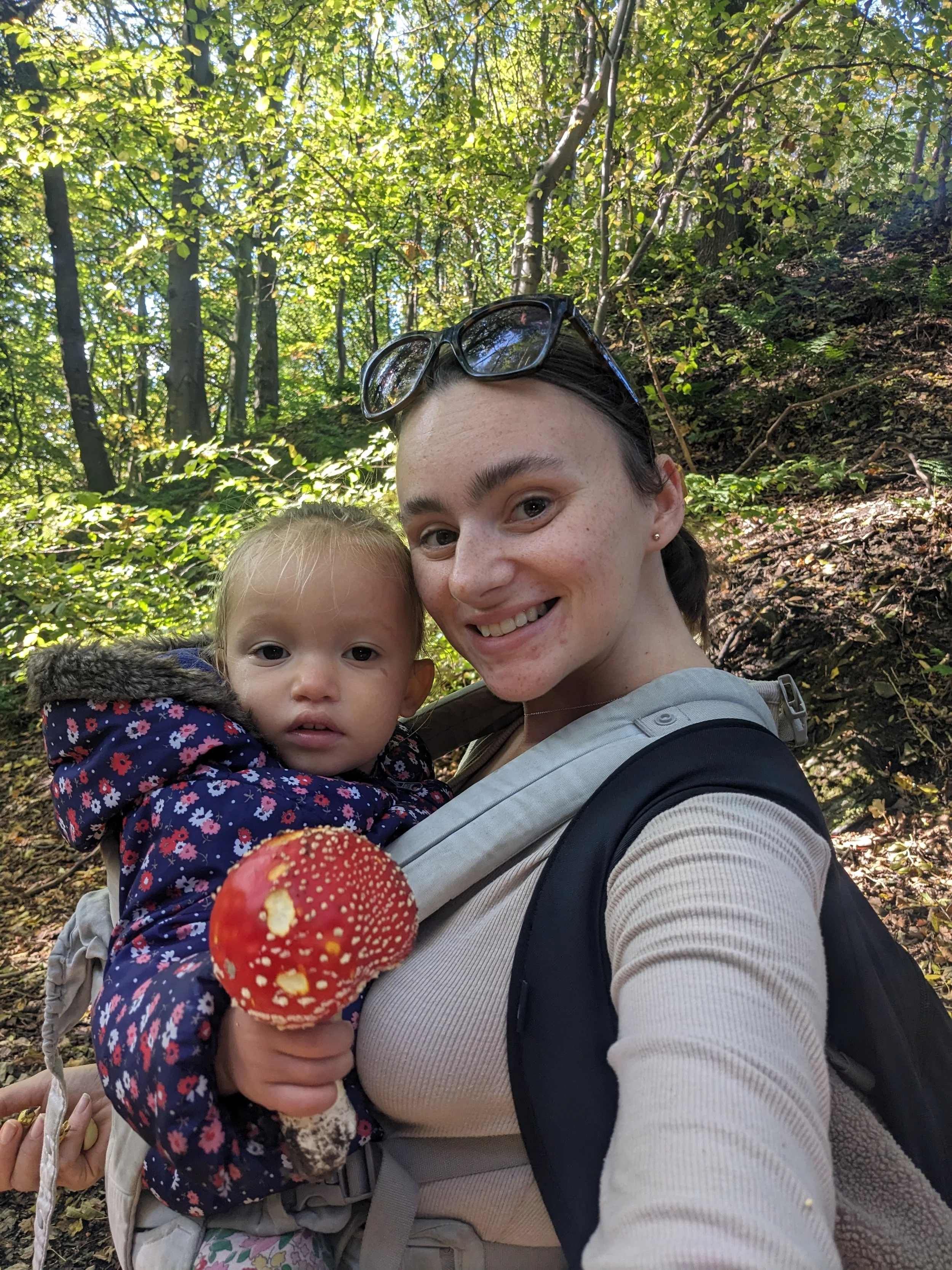 A young woman with sunglasses on her head holding a young girl with blonde hair, both smiling. The girl is wearing a floral jacket. The woman is holding a bright red mushroom with white spots in her hand. They are in a forest with green leaves and su
