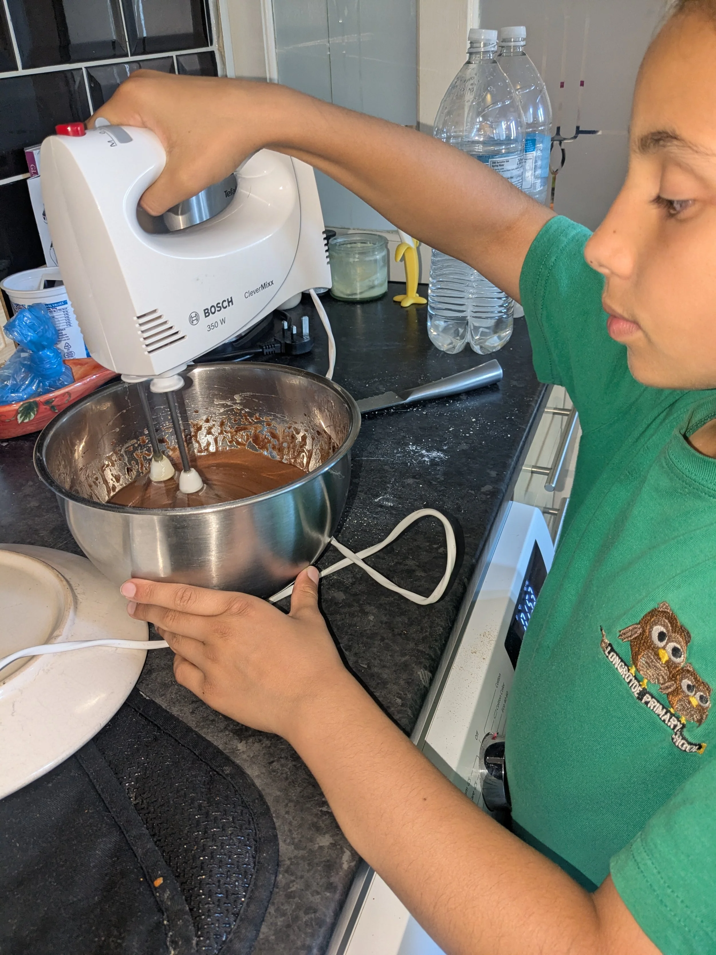 A person using a handheld electric mixer to stir chocolate batter in a metal bowl in a kitchen.