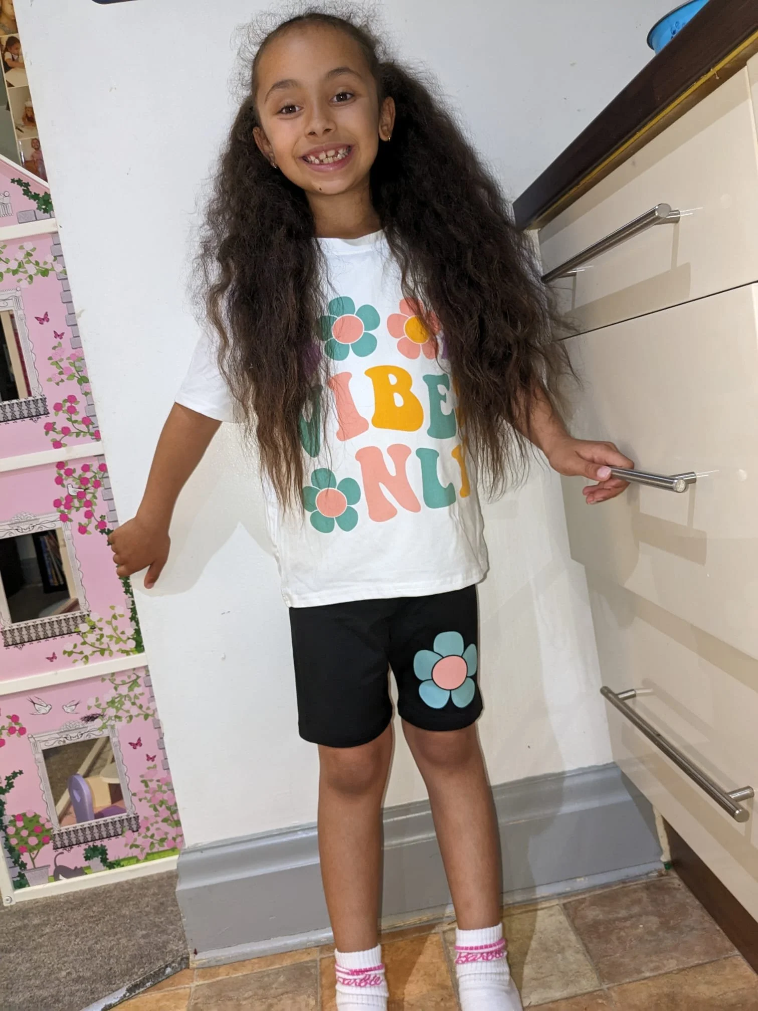 A young girl with long curly hair smiling, standing indoors near a white wall and a beige dresser, wearing a white t-shirt with colorful flower and letter print, black shorts with a flower graphic, pink and white socks, and white shoes.