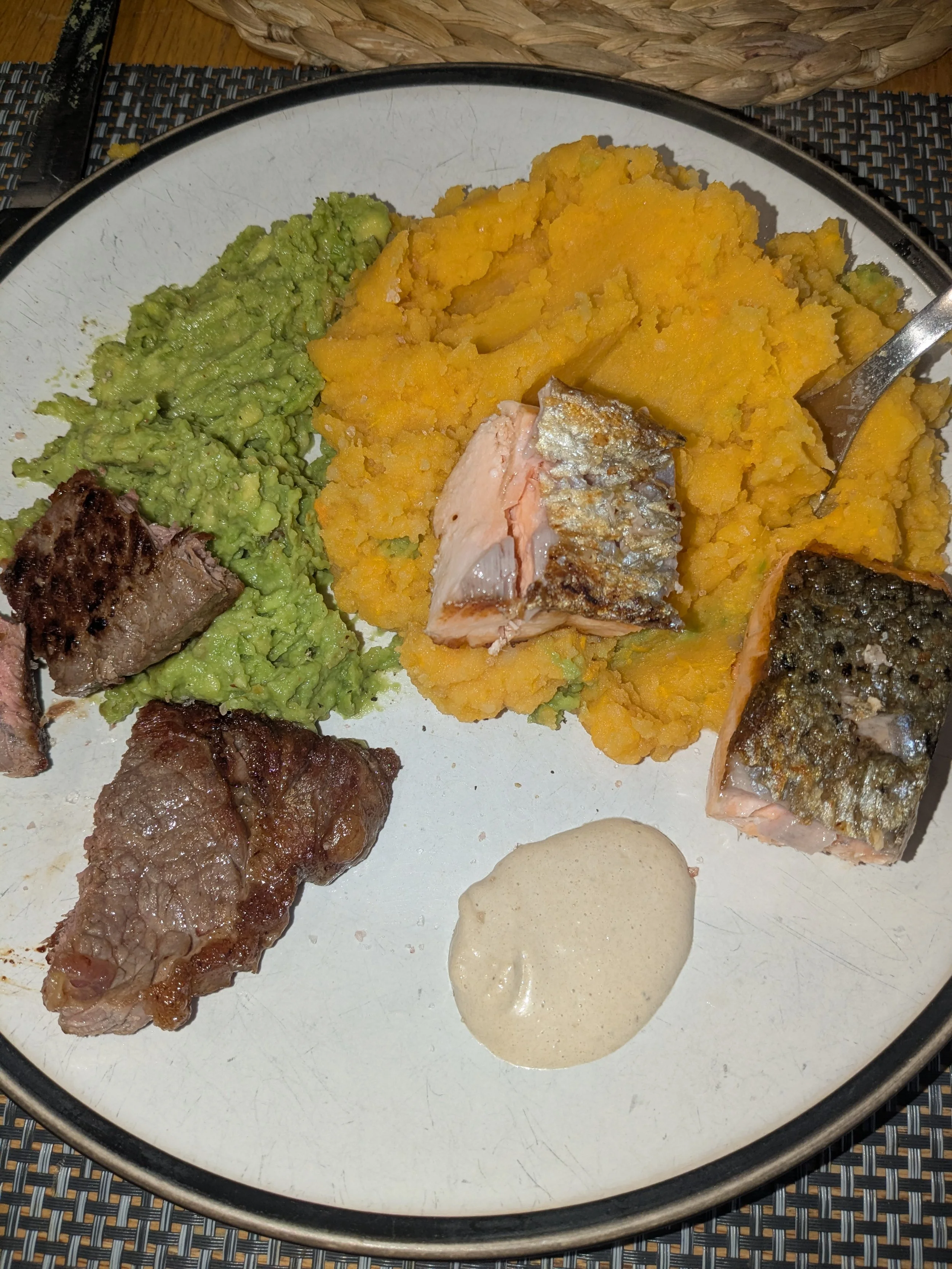 A plate of cooked food including mashed yellow squash, guacamole, cooked fish with skin, cooked steak, and a dollop of white sauce or dip.