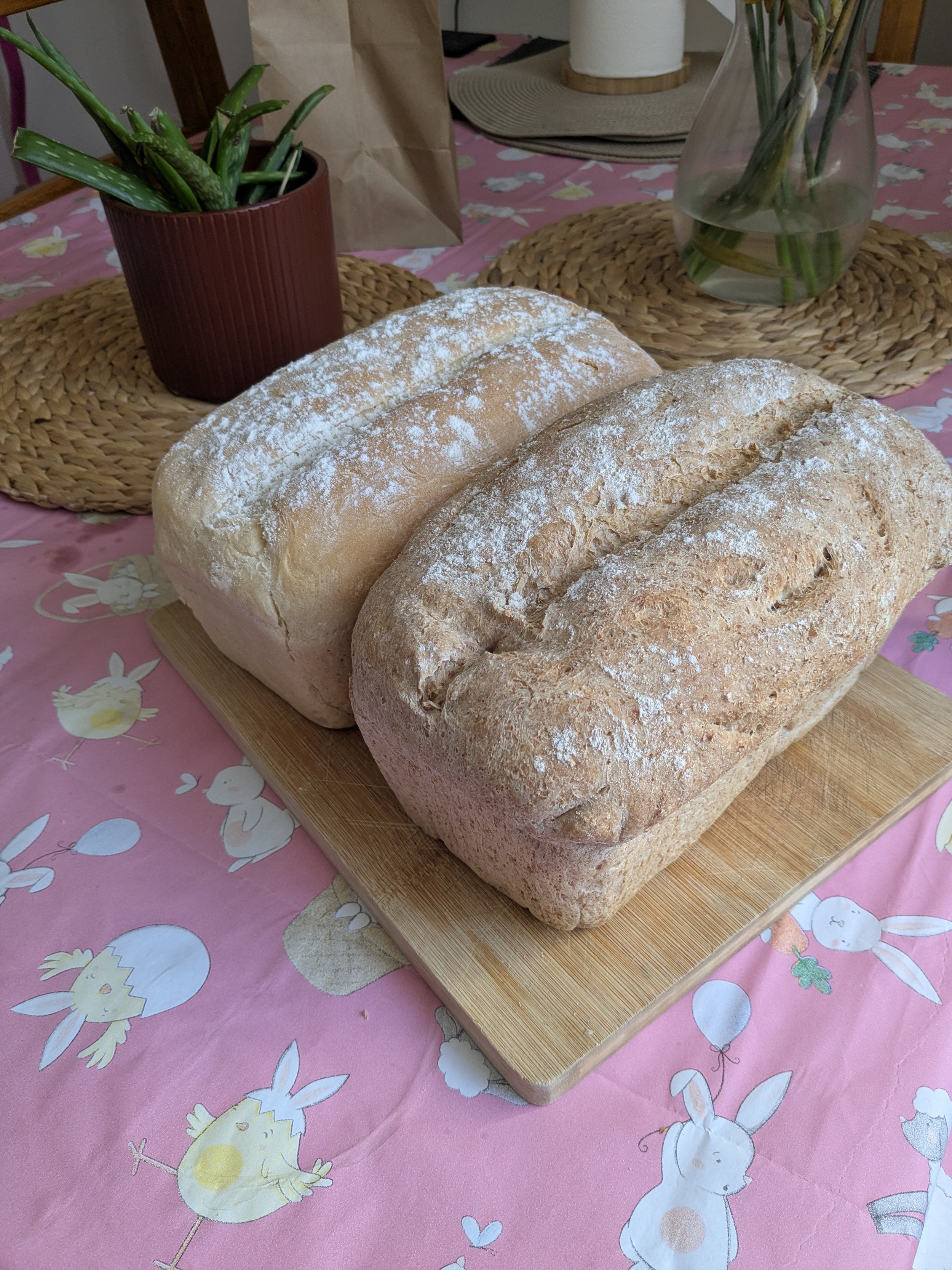 Two loaves of dusted bread on a wooden cutting board, with a pink tablecloth decorated with cartoon rabbits and other animals, and a potted aloe vera plant and a vase with flowers in the background.