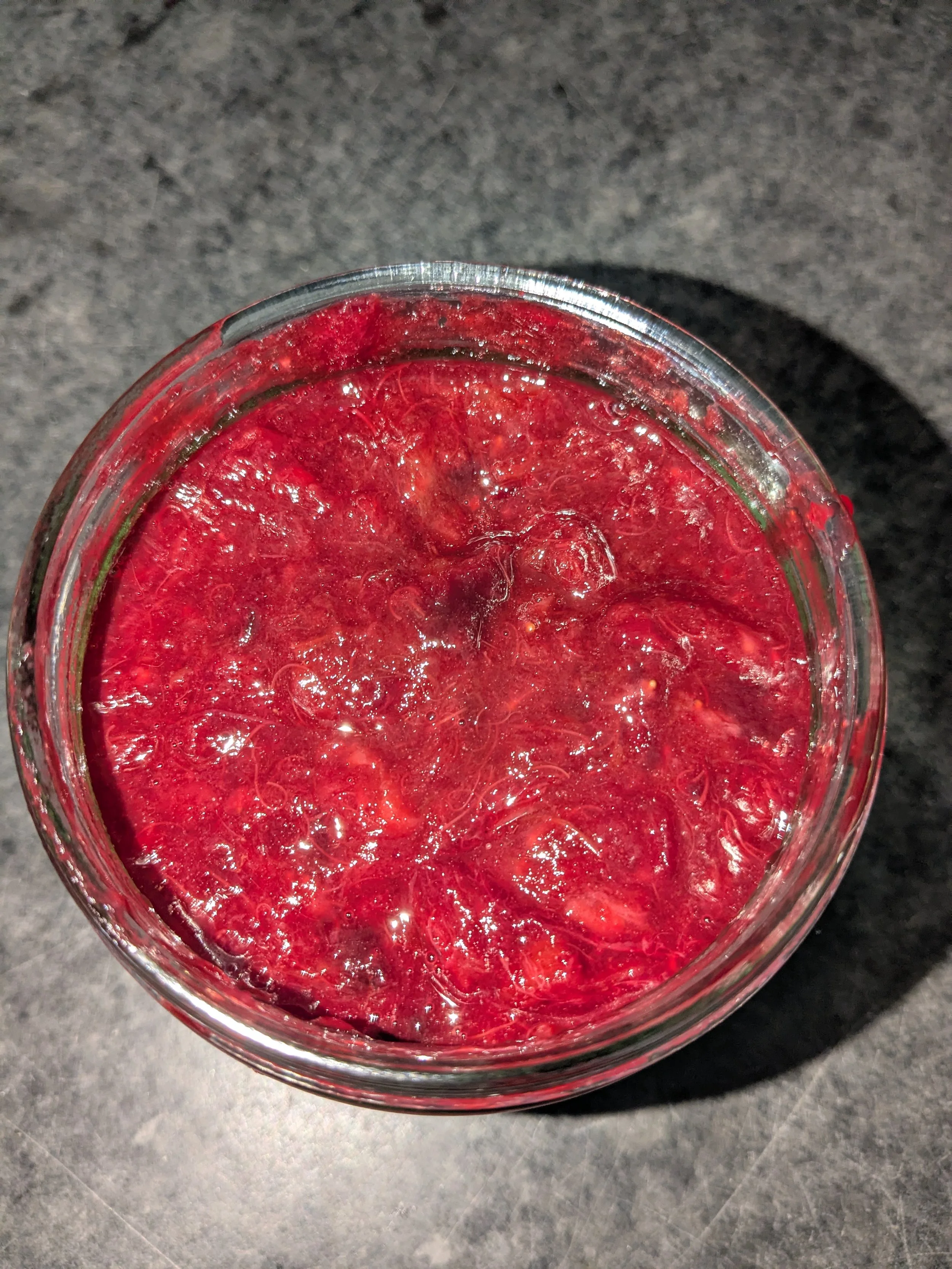A top view of a clear glass jar filled with a red, frozen beverage containing visible ice and fruit pieces, placed on a dark gray countertop.
