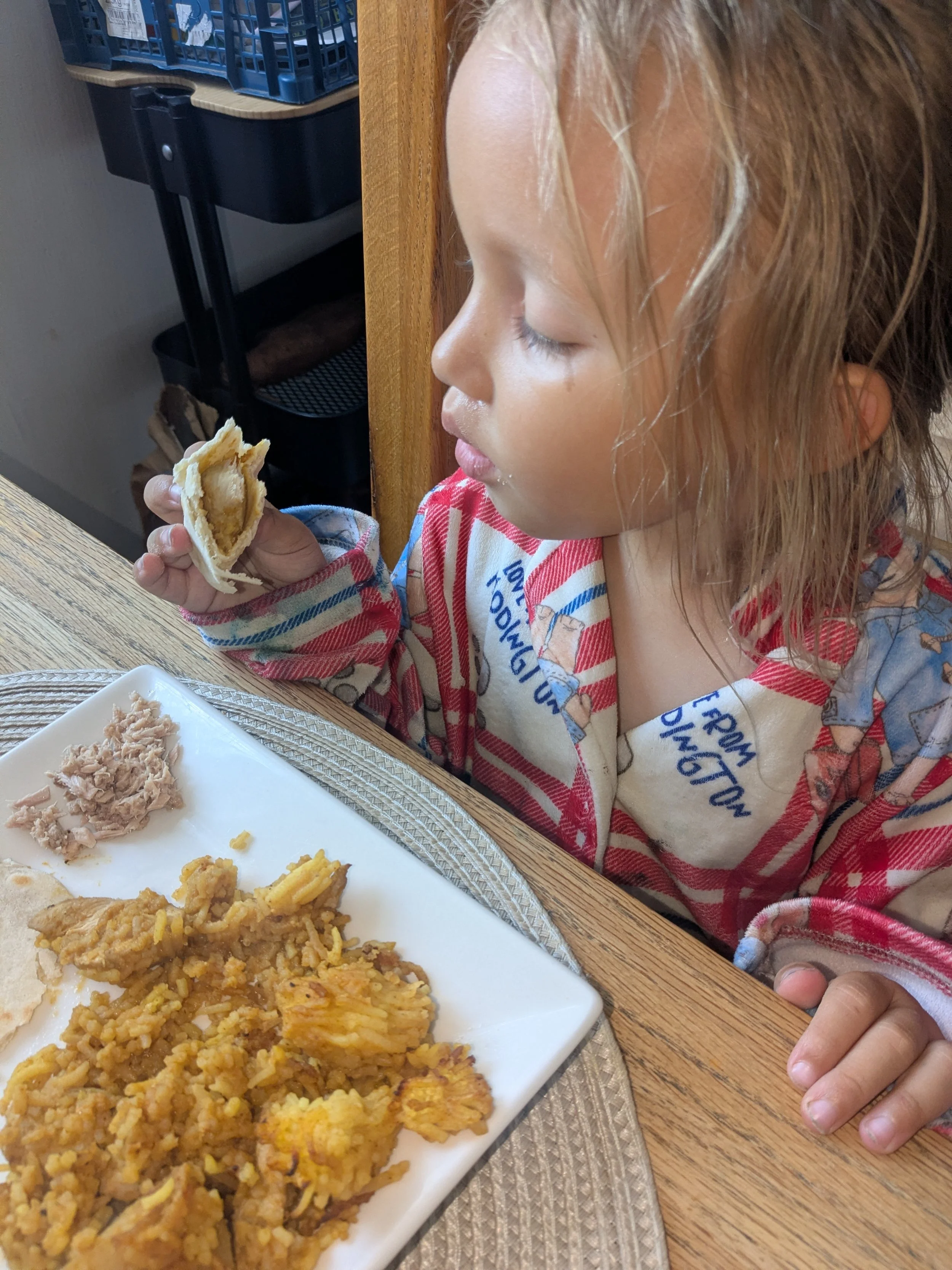 A young girl with wet hair sits at a wooden table, holding a half-eaten taco. In front of her is a white plate with fried rice, corn, and meat. The background includes a black shelving unit and a blue plastic container.