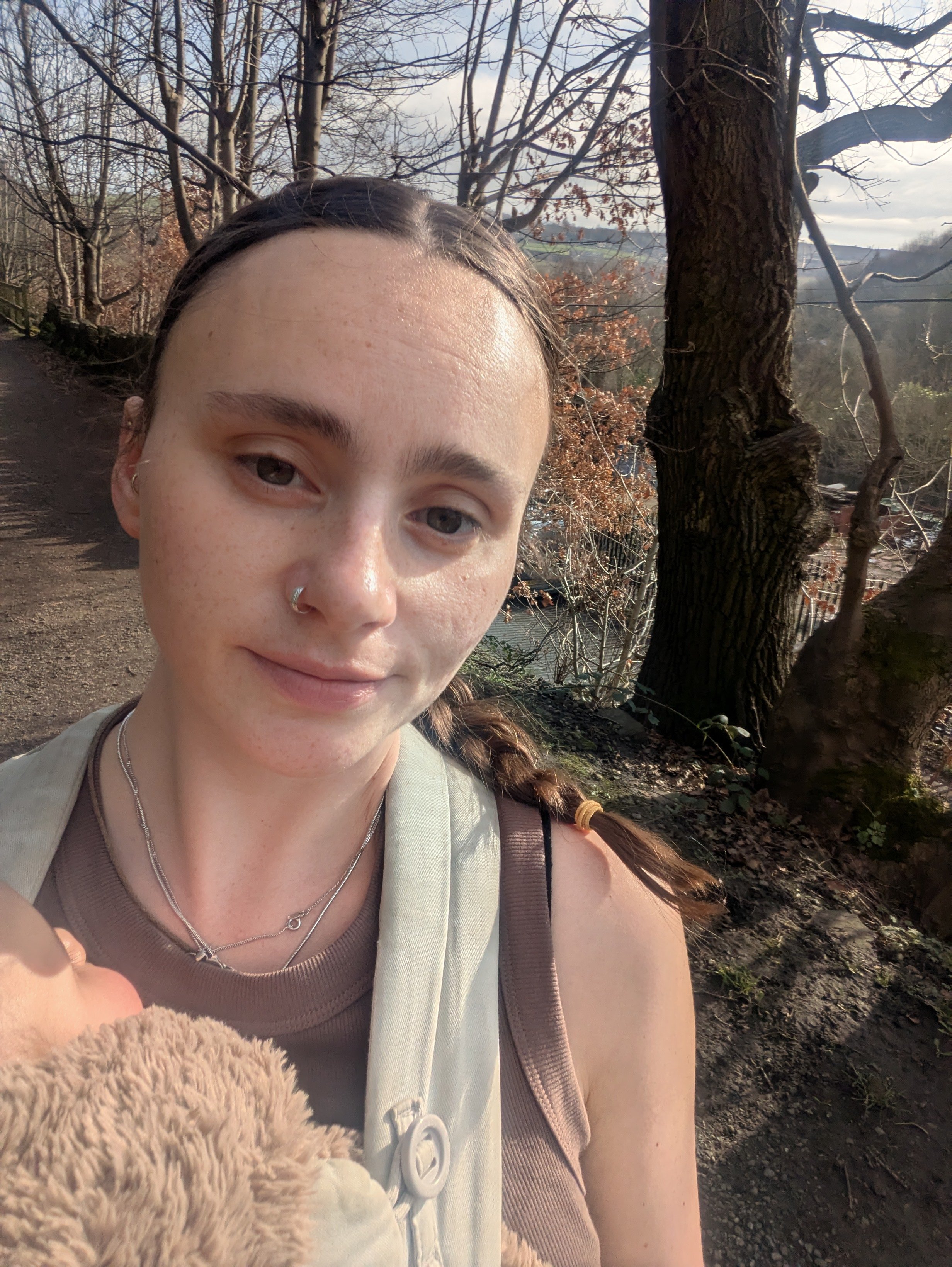 A woman with freckles and multiple necklaces taking a selfie outdoors on a trail, with a large twisted tree and leafless branches in the background.