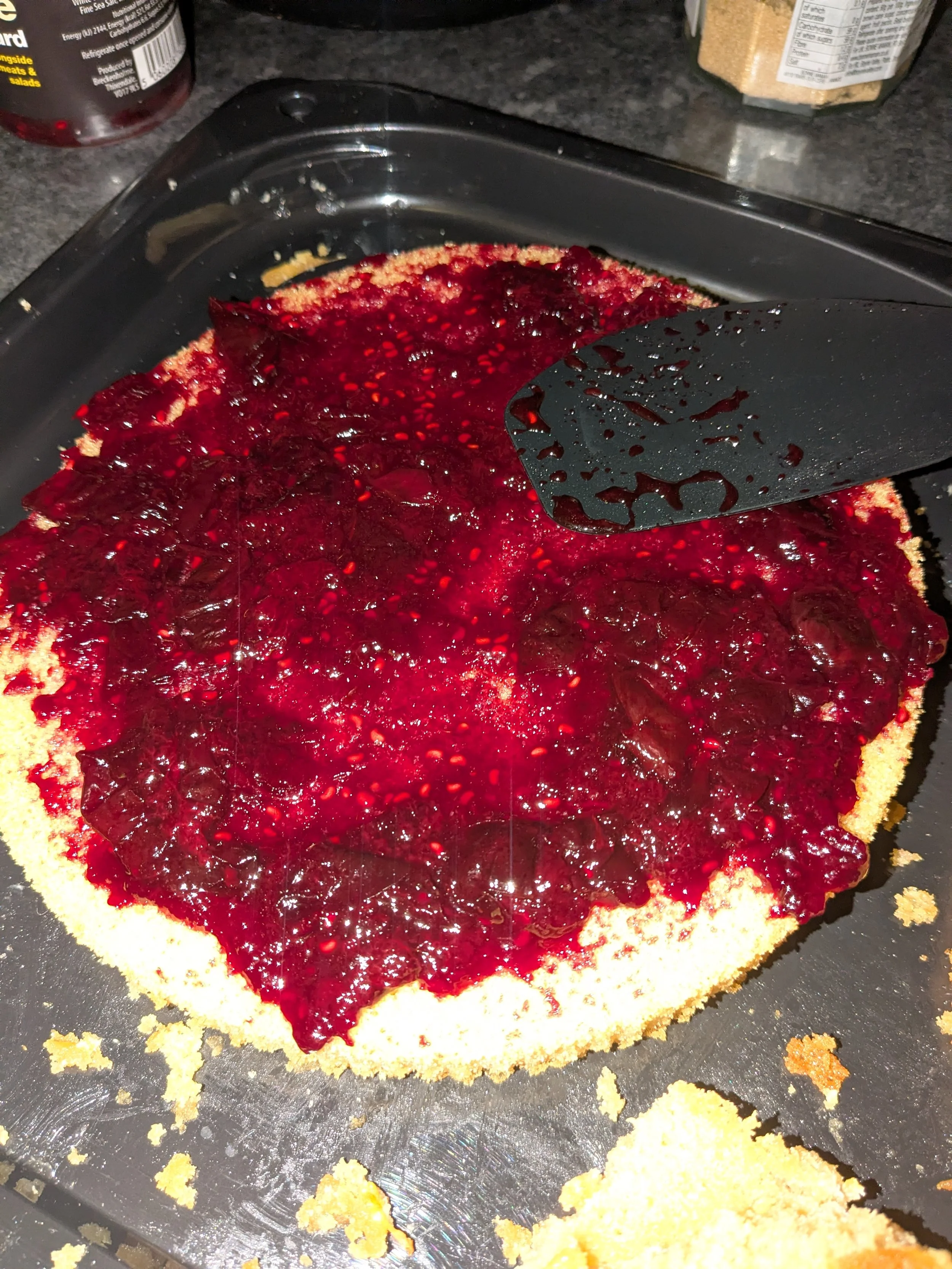 A slice of cheesecake topped with red berry compote on a black tray, with crumbs and crumbs scattered around.
