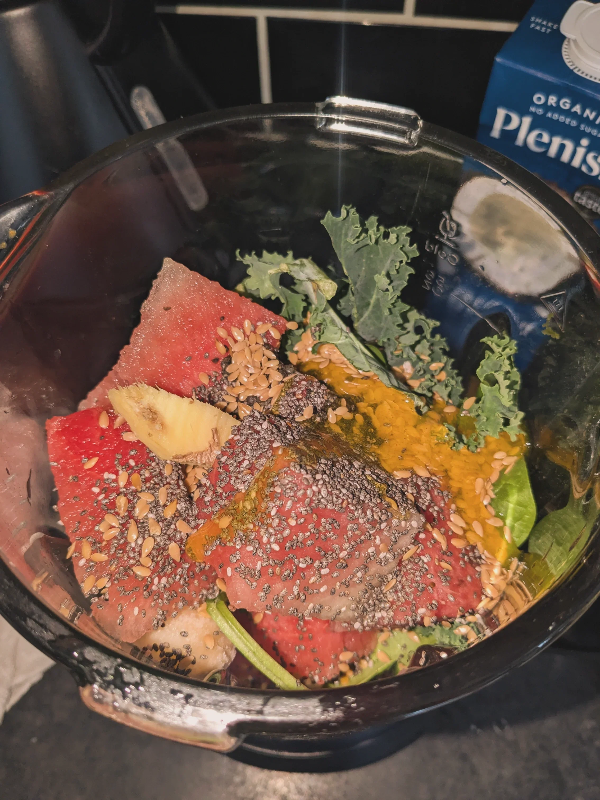 An air fryer containing sliced watermelon, banana, kale leaves, and topped with seeds.