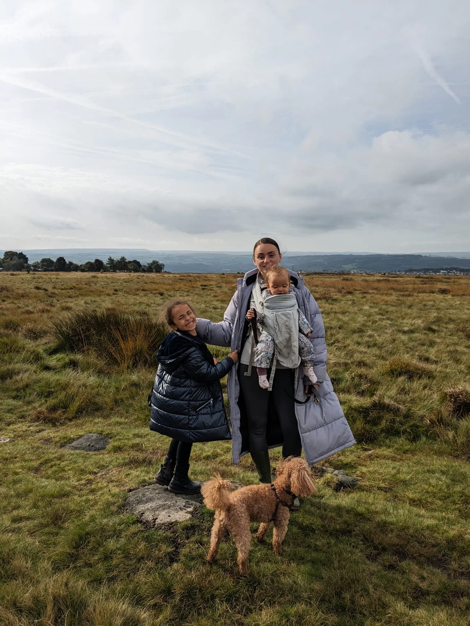 Three children with a woman holding a baby in a grassy outdoor field, with a large brown dog standing in the foreground and a cloudy sky in the background.