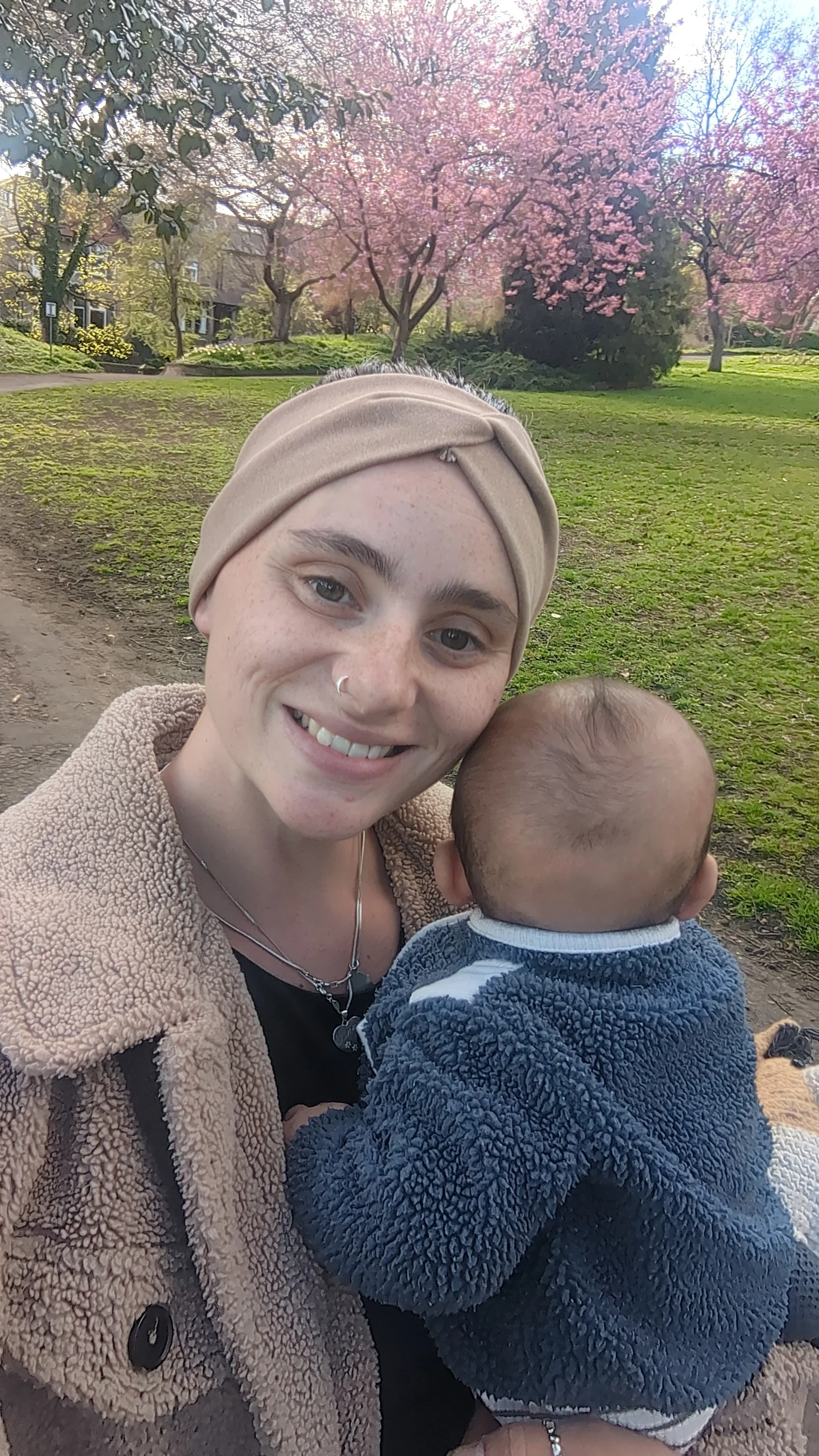 A woman smiling and holding a young child outdoors in a park with trees and blooming pink flowers in the background.