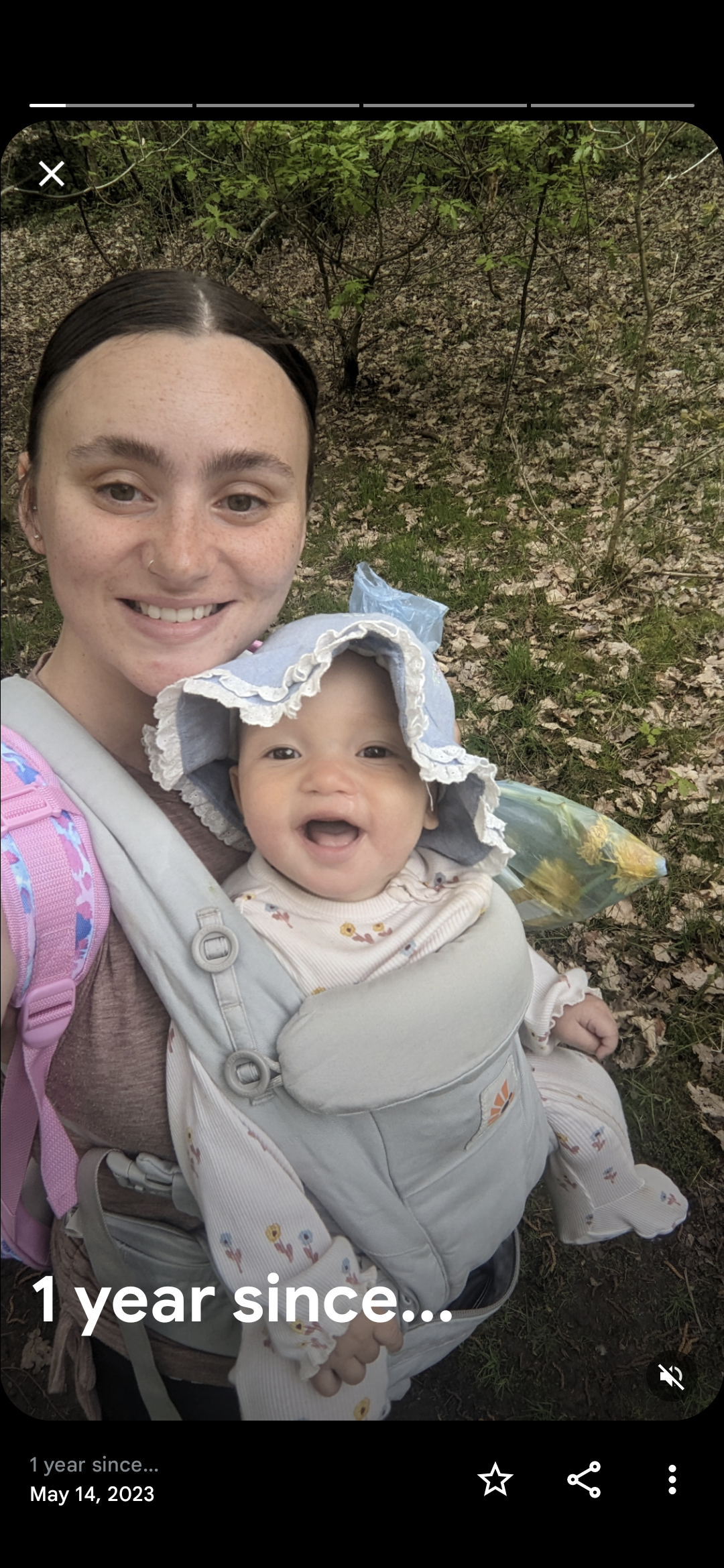 A woman with dark hair in a baby carrier holding a smiling baby girl wearing a sun hat, outdoors with green foliage and fallen leaves in the background.
