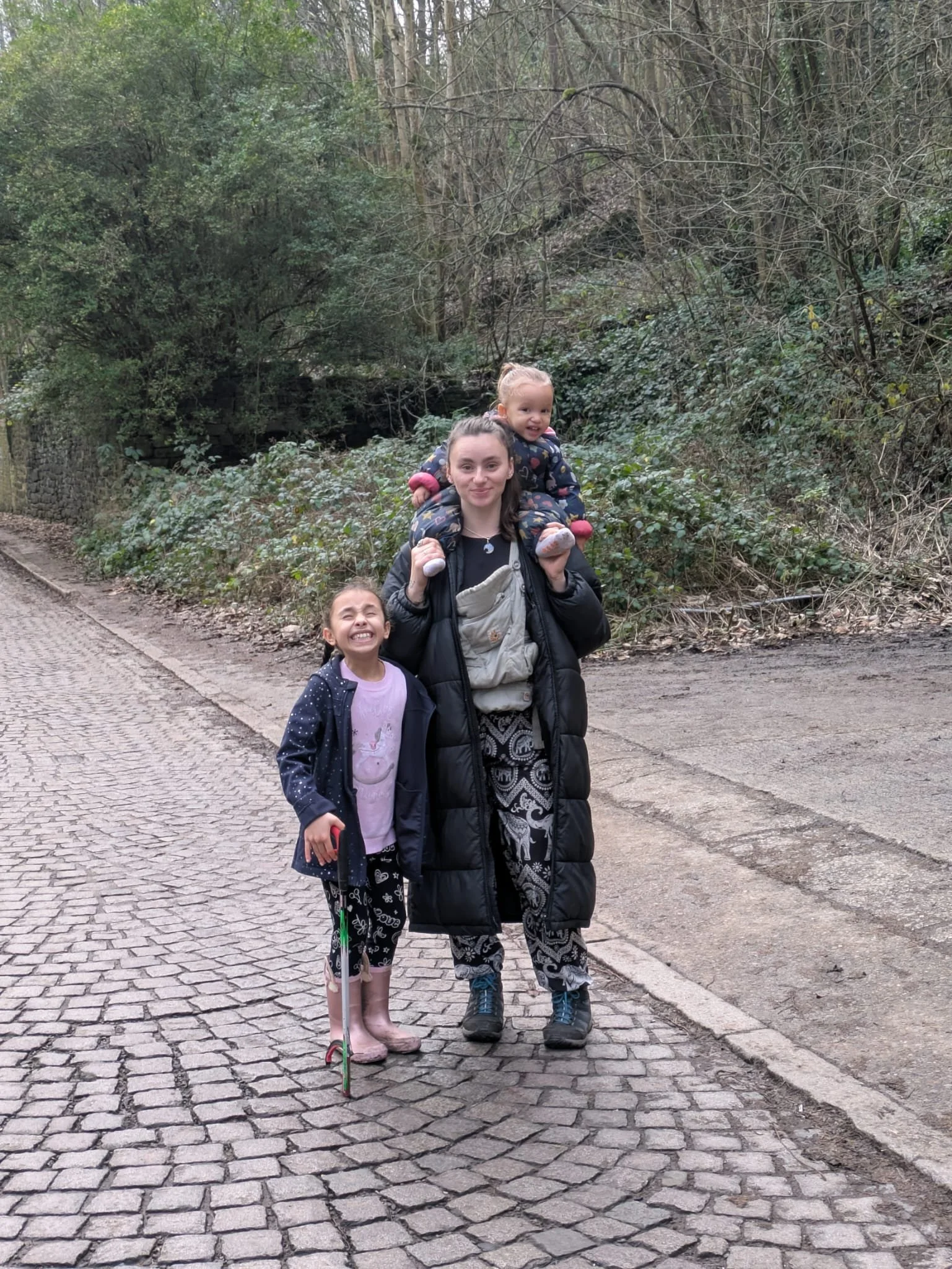 A woman with two young girls on a cobblestone path in a wooded area. The woman carries a girl on her shoulders, while the other girl stands beside her. The girl on the shoulders is smiling while the other girl is smiling with her eyes closed. The bac