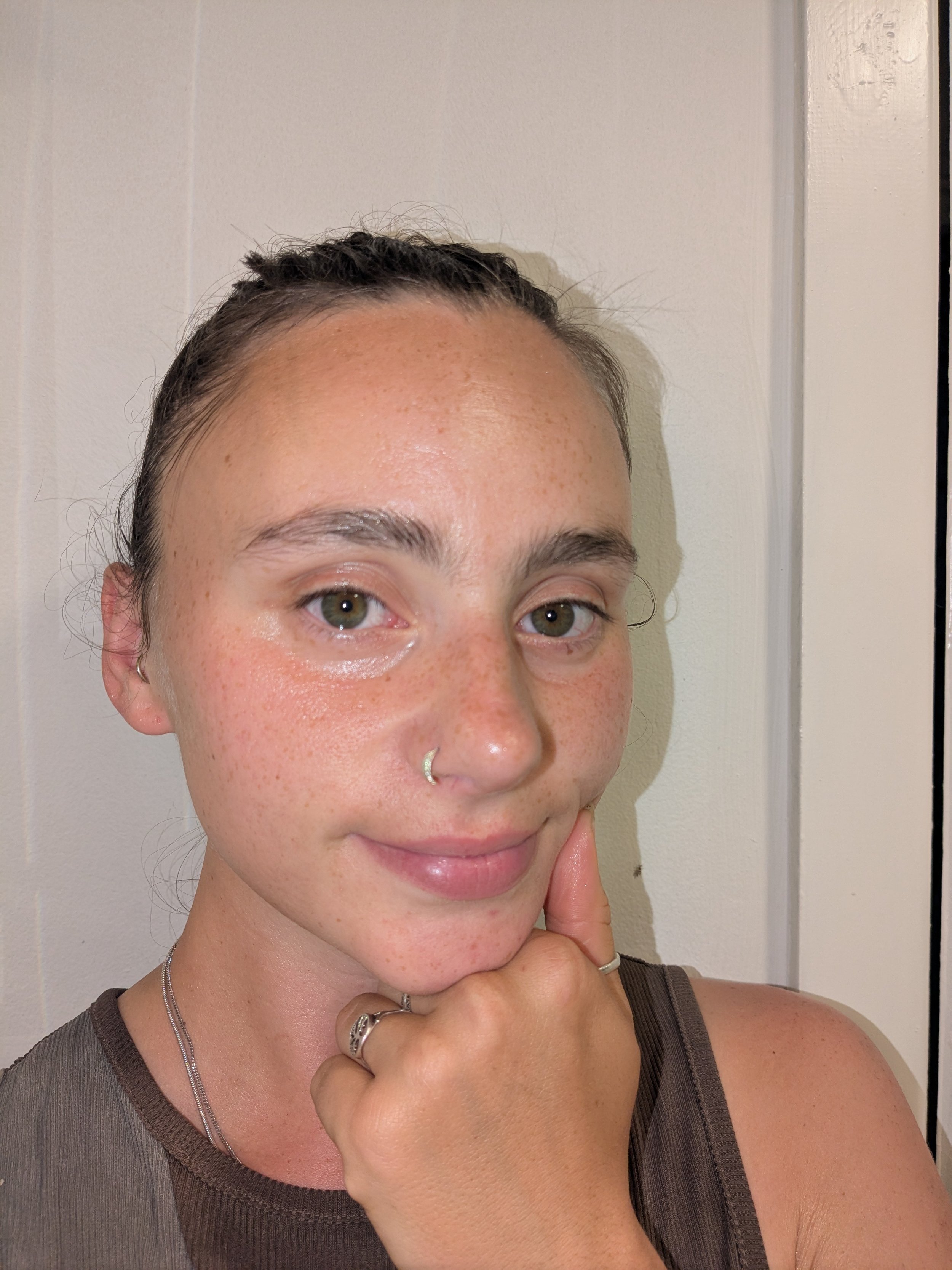 Close-up of a woman with short hair, freckles, and green eyes, smiling and holding her chin with her hand, wearing a tank top and silver jewelry against a white wall background.
